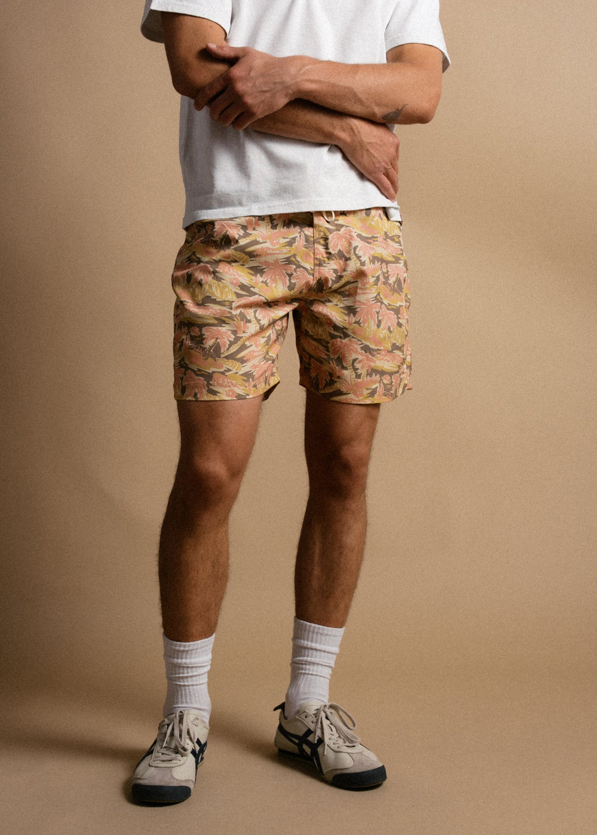 Man in white shirt and floral shorts stands against a beige background, wearing sneakers and socks, arms crossed casually.