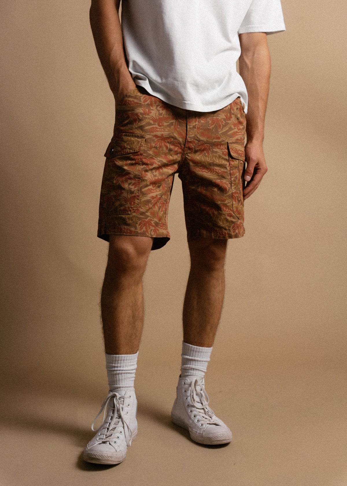 Man in a white t-shirt, floral cargo shorts, and white sneakers stands against a beige background.