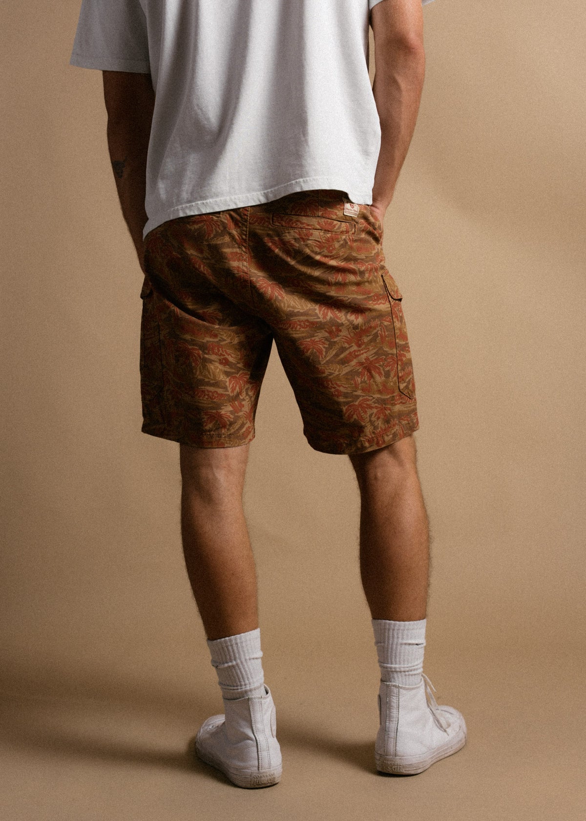 Man in white shirt and patterned shorts stands against a beige background, wearing white socks and sneakers.