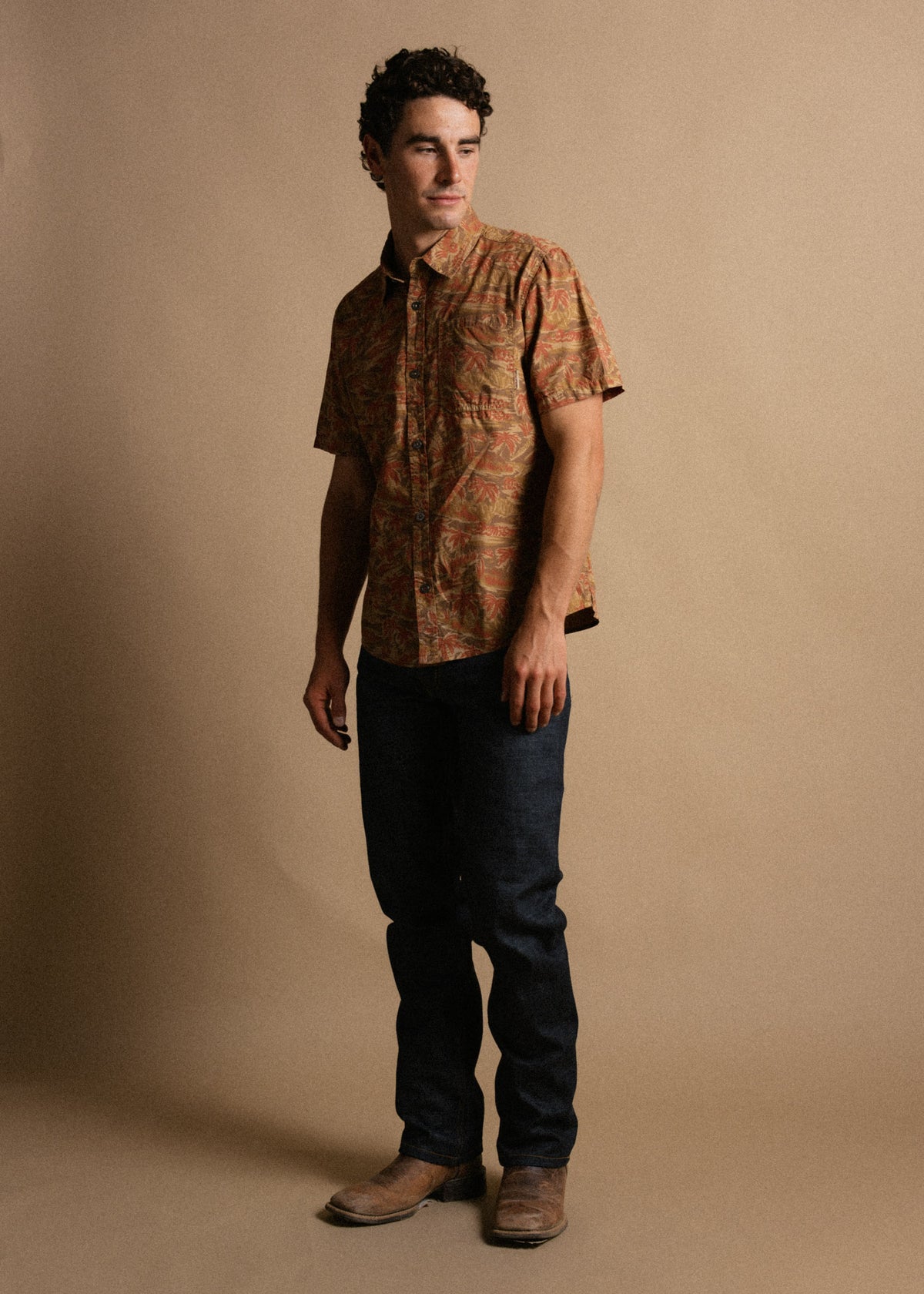 Man in patterned shirt, jeans, and boots stands against a beige background, looking to the side with a thoughtful expression.