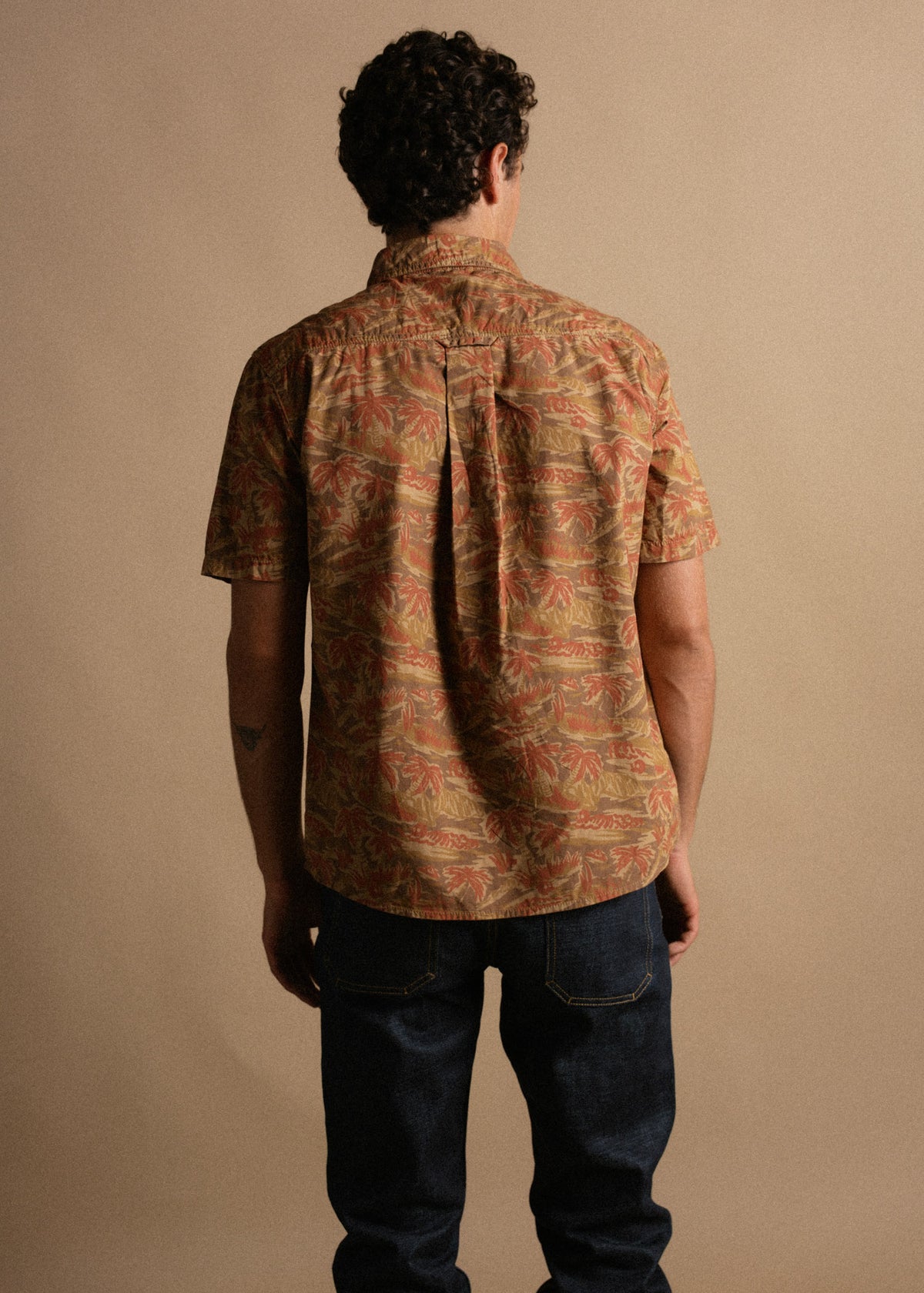 Man in floral shirt and jeans stands with back to camera against a neutral background, showcasing casual fashion style.