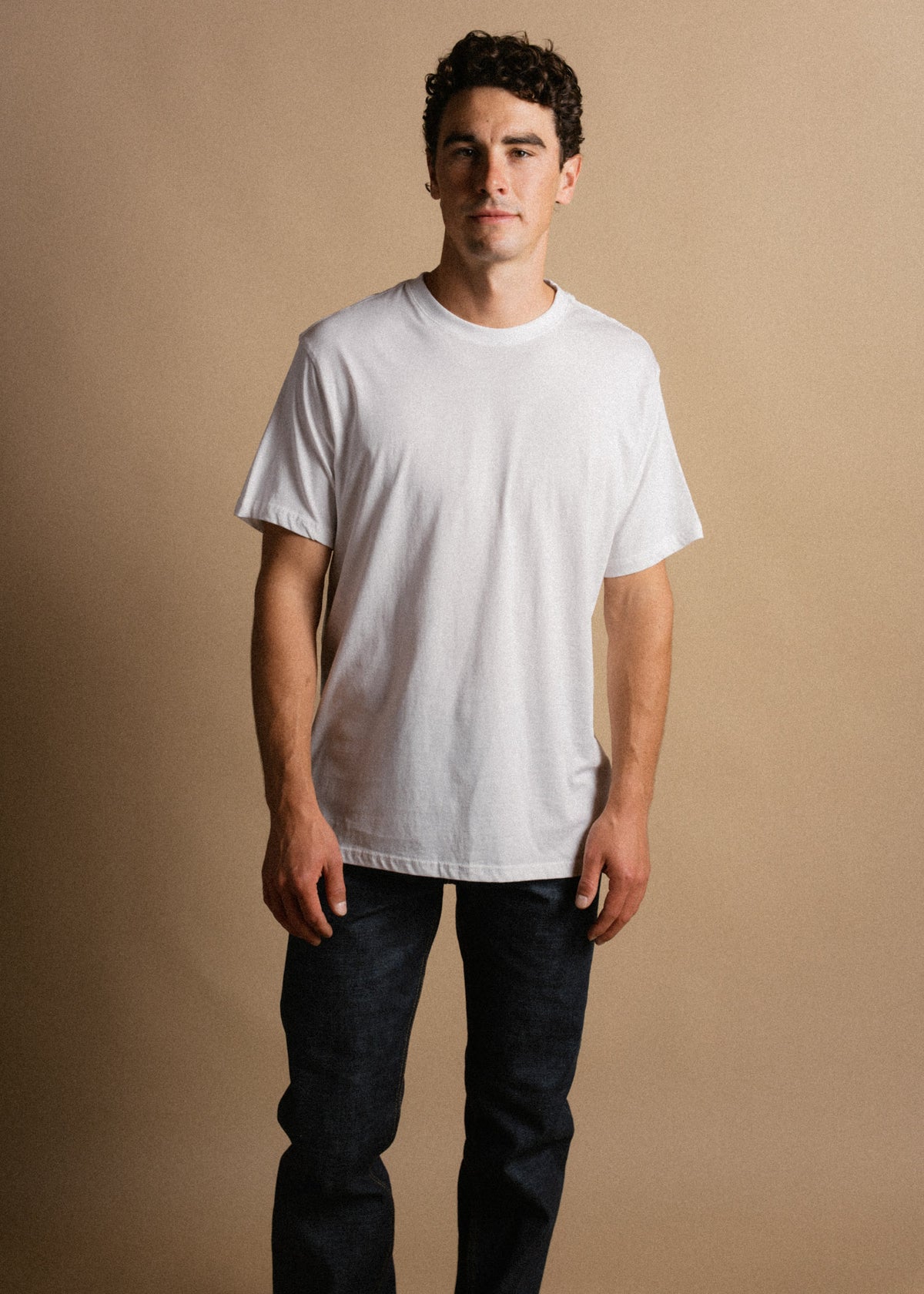 Man in a white T-shirt and dark jeans stands against a beige background, looking confidently at the camera.