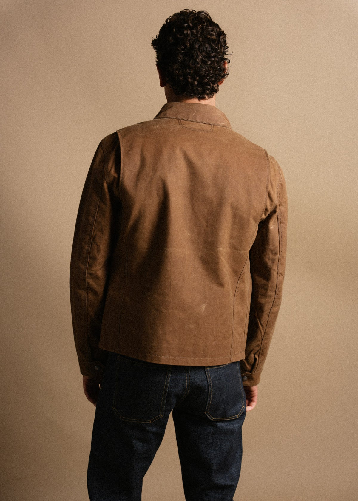 Man in brown leather jacket and dark jeans stands with back to camera against a beige background, showcasing casual style.