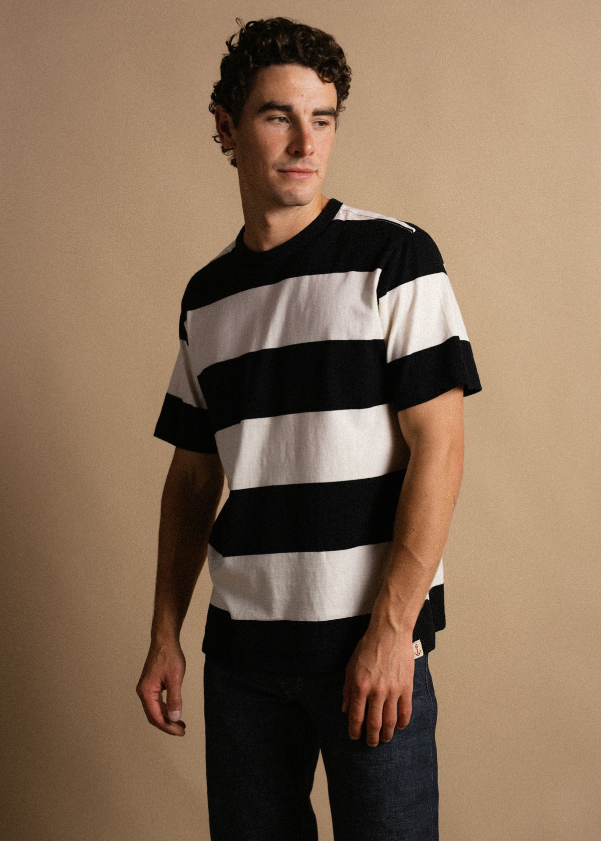 Man in a black and white striped shirt stands against a beige background, looking to the side with a relaxed expression.
