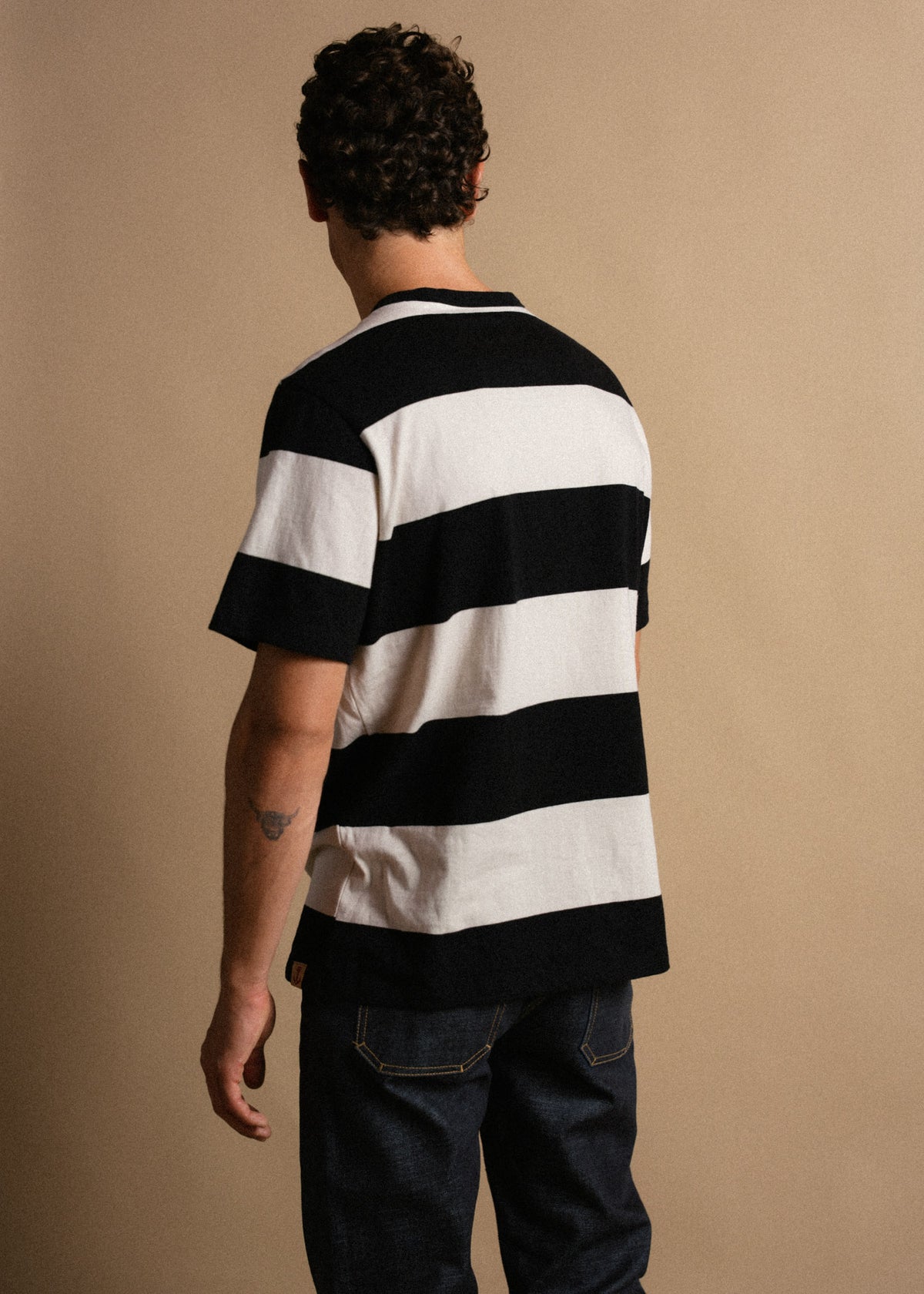 Man in a black and white striped shirt and jeans stands with his back to the camera against a plain beige background.