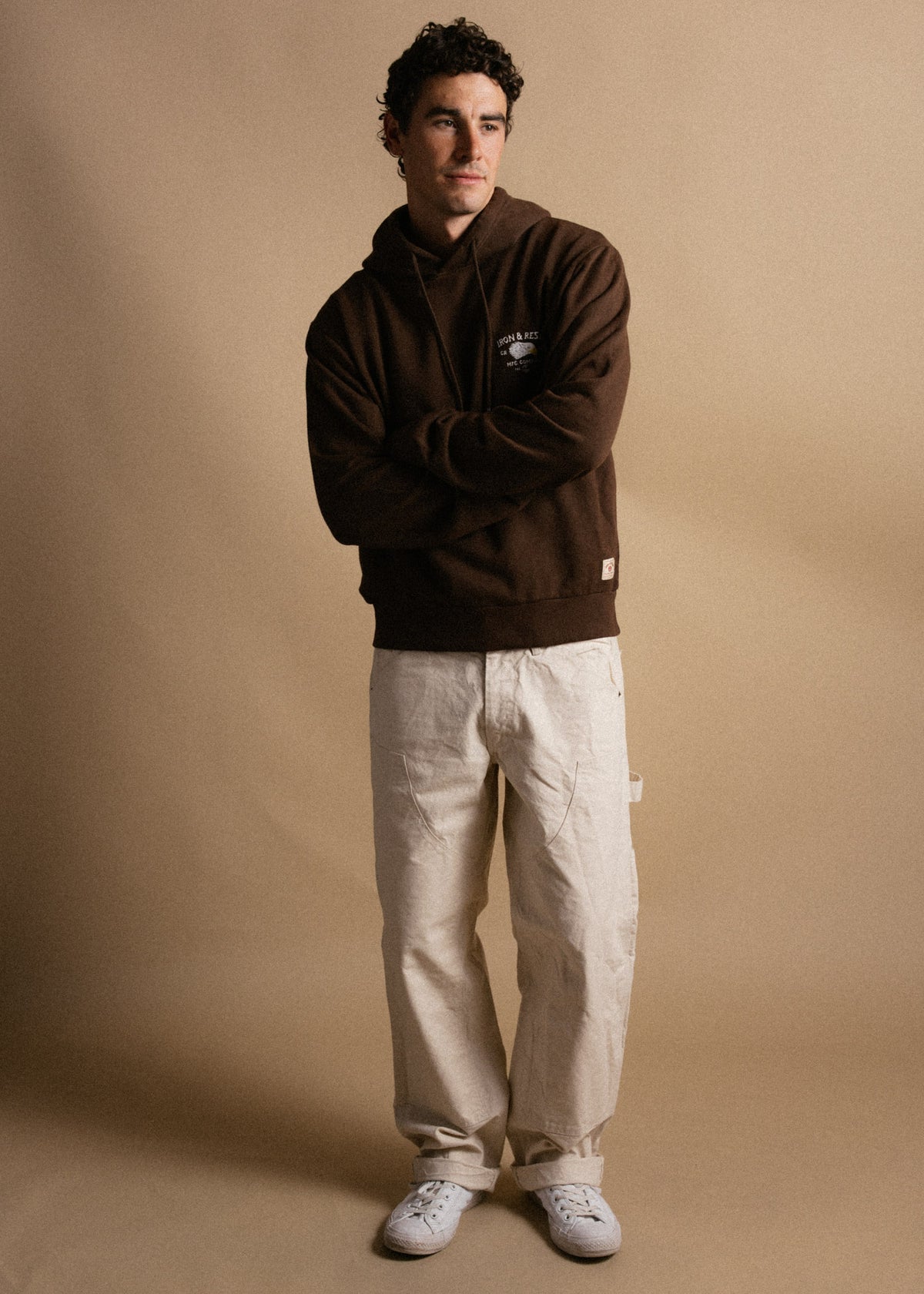 Man in brown hoodie and beige pants stands against a tan backdrop, arms crossed, looking to the side, wearing white sneakers.