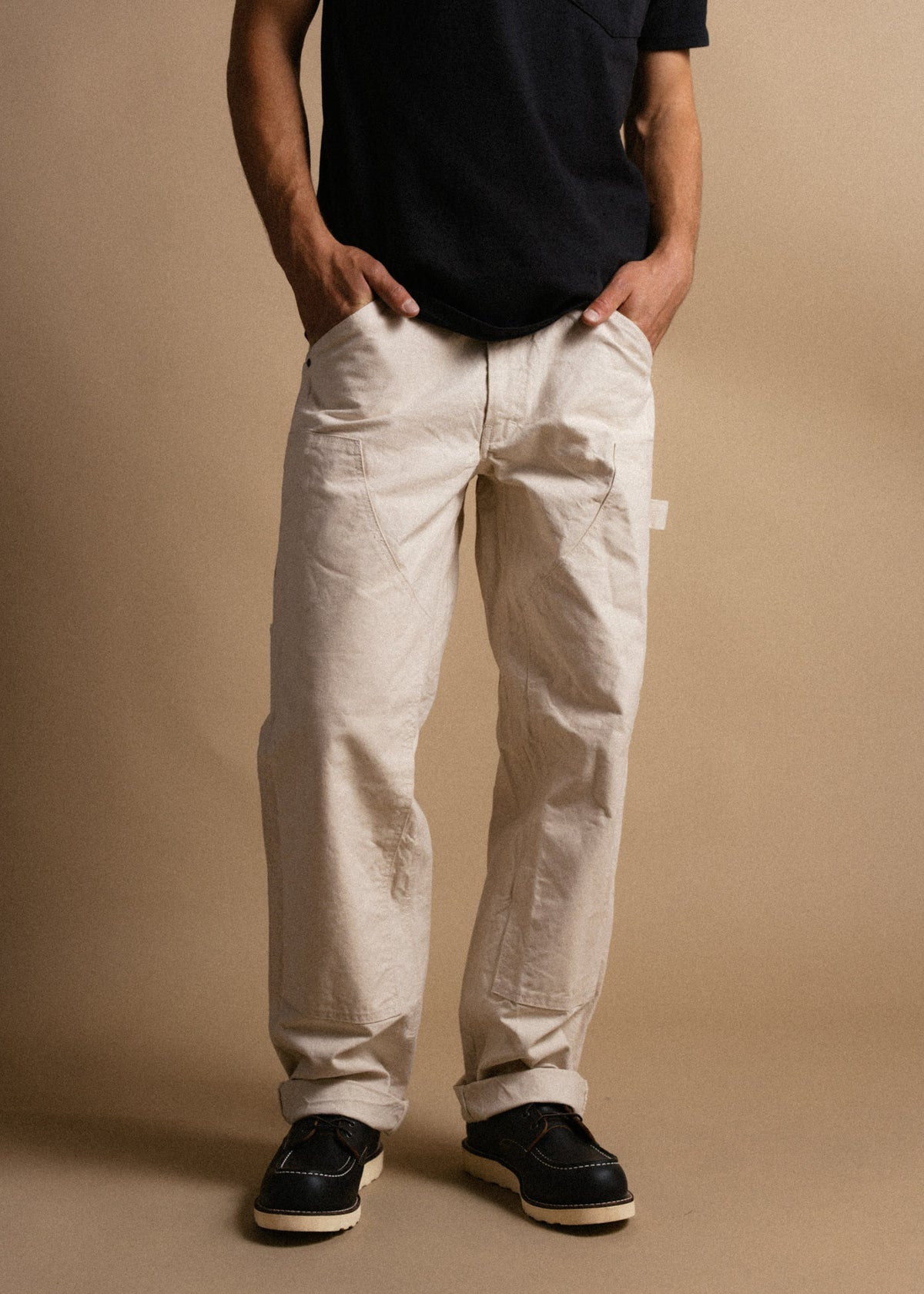 Man in black shirt and beige pants stands with hands in pockets, wearing black shoes, against a plain tan background.