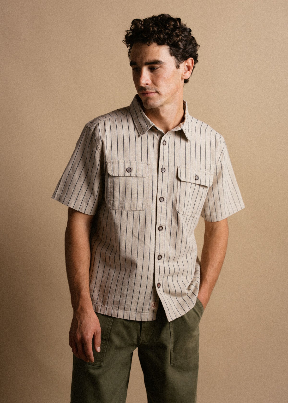 Man in a striped short-sleeve shirt and green pants stands against a beige background, looking to the side with a relaxed pose.