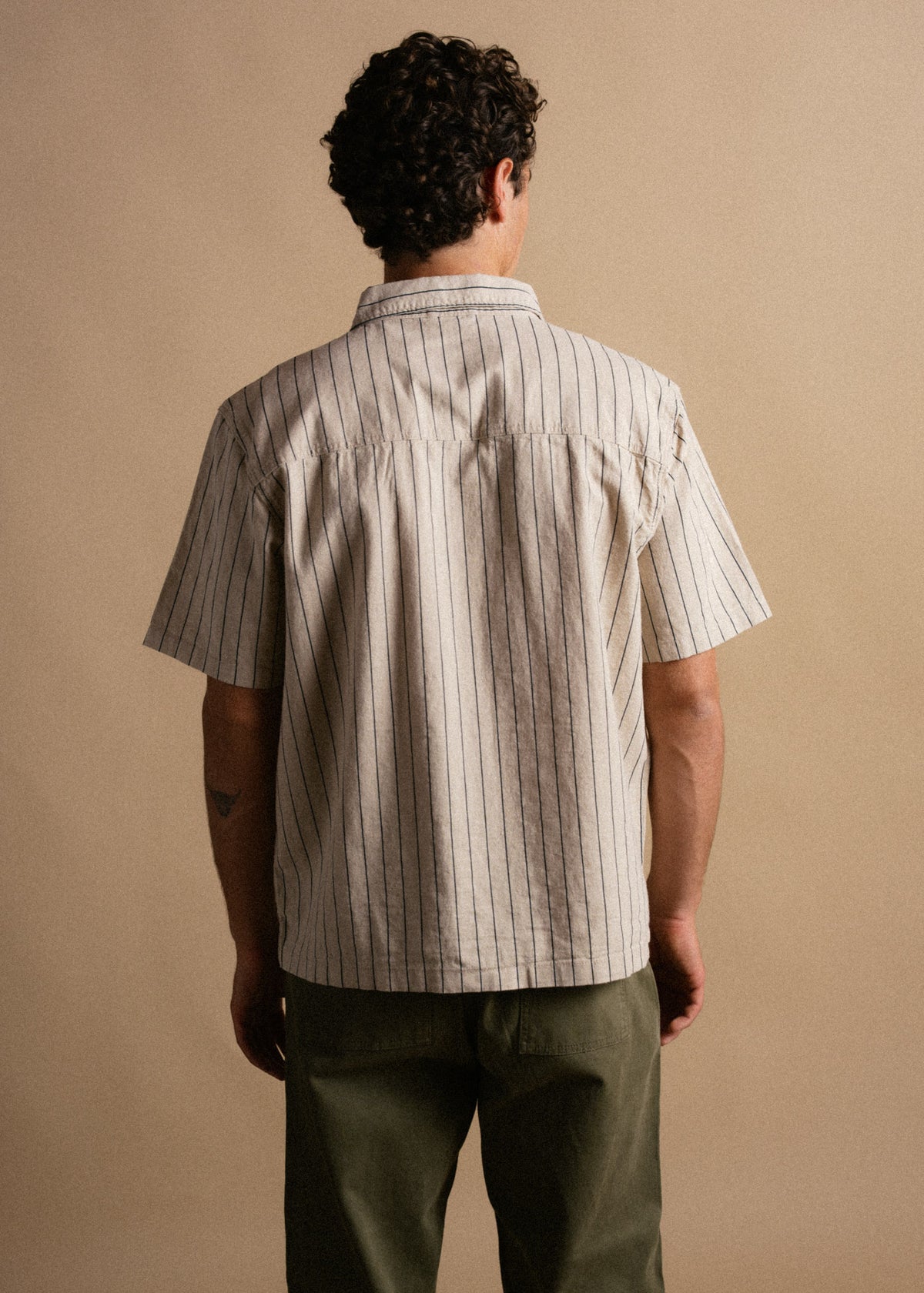 Man in a striped short-sleeve shirt and green pants stands with his back to the camera against a beige background.