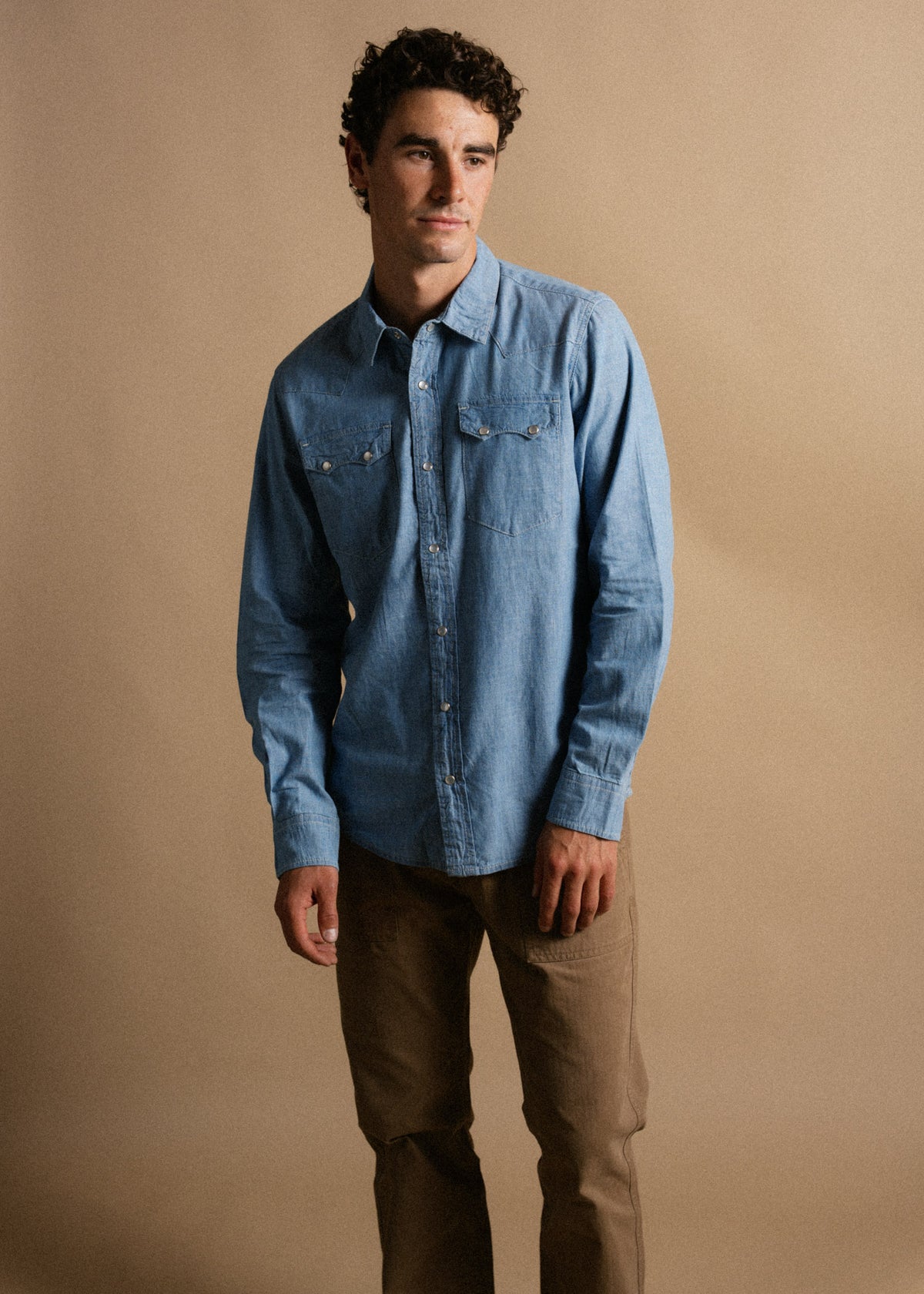 A man in a blue denim shirt and brown pants stands against a beige background, looking to the side with a thoughtful expression.