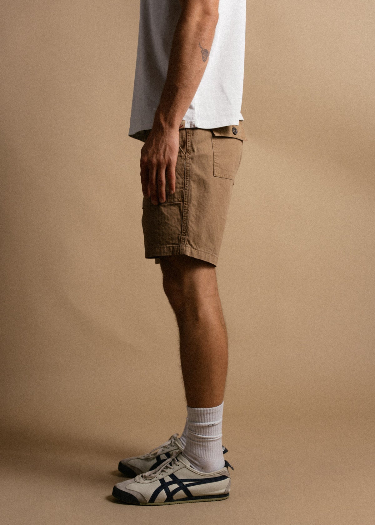 Man in white shirt, beige shorts, and sneakers stands sideways against a plain background, showcasing casual summer style.