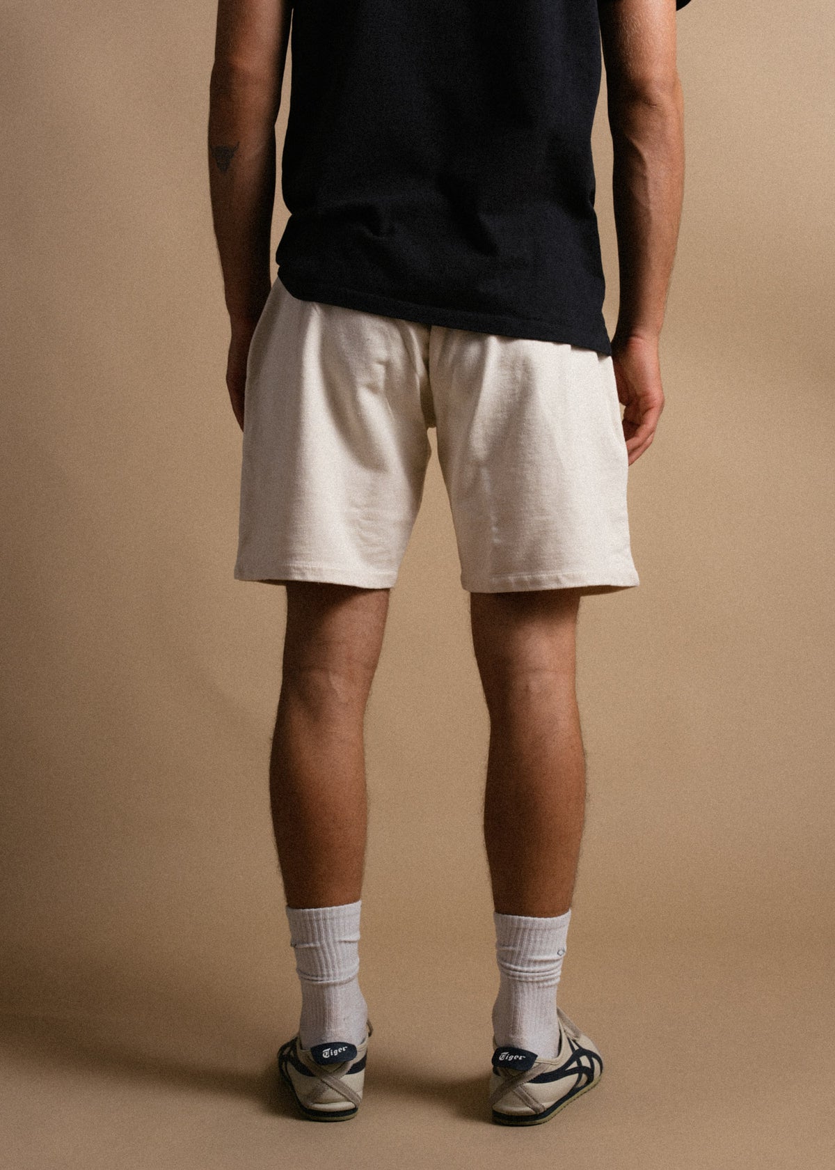 Man in black shirt and beige shorts stands facing away, wearing white socks and sneakers, against a plain beige background.