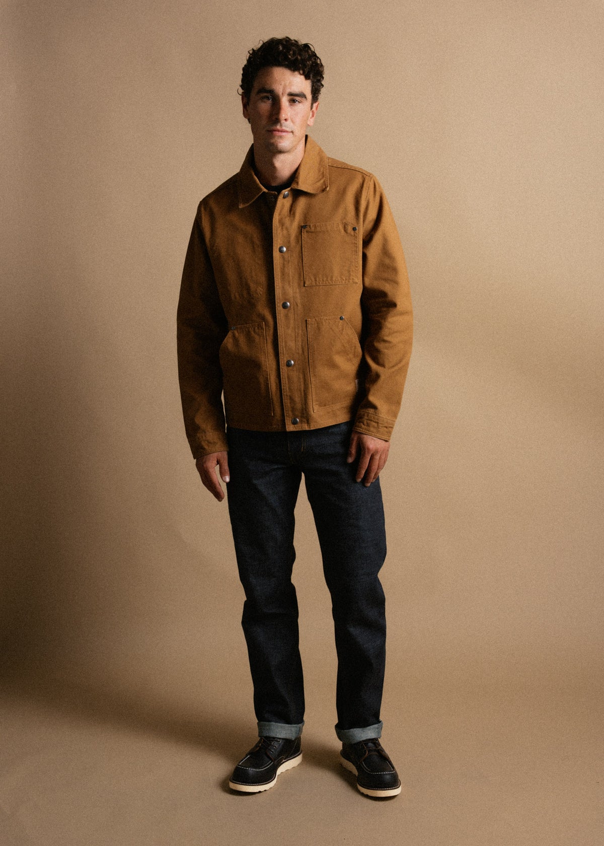 Man in brown jacket and dark jeans stands against a beige background, looking at the camera with a neutral expression.