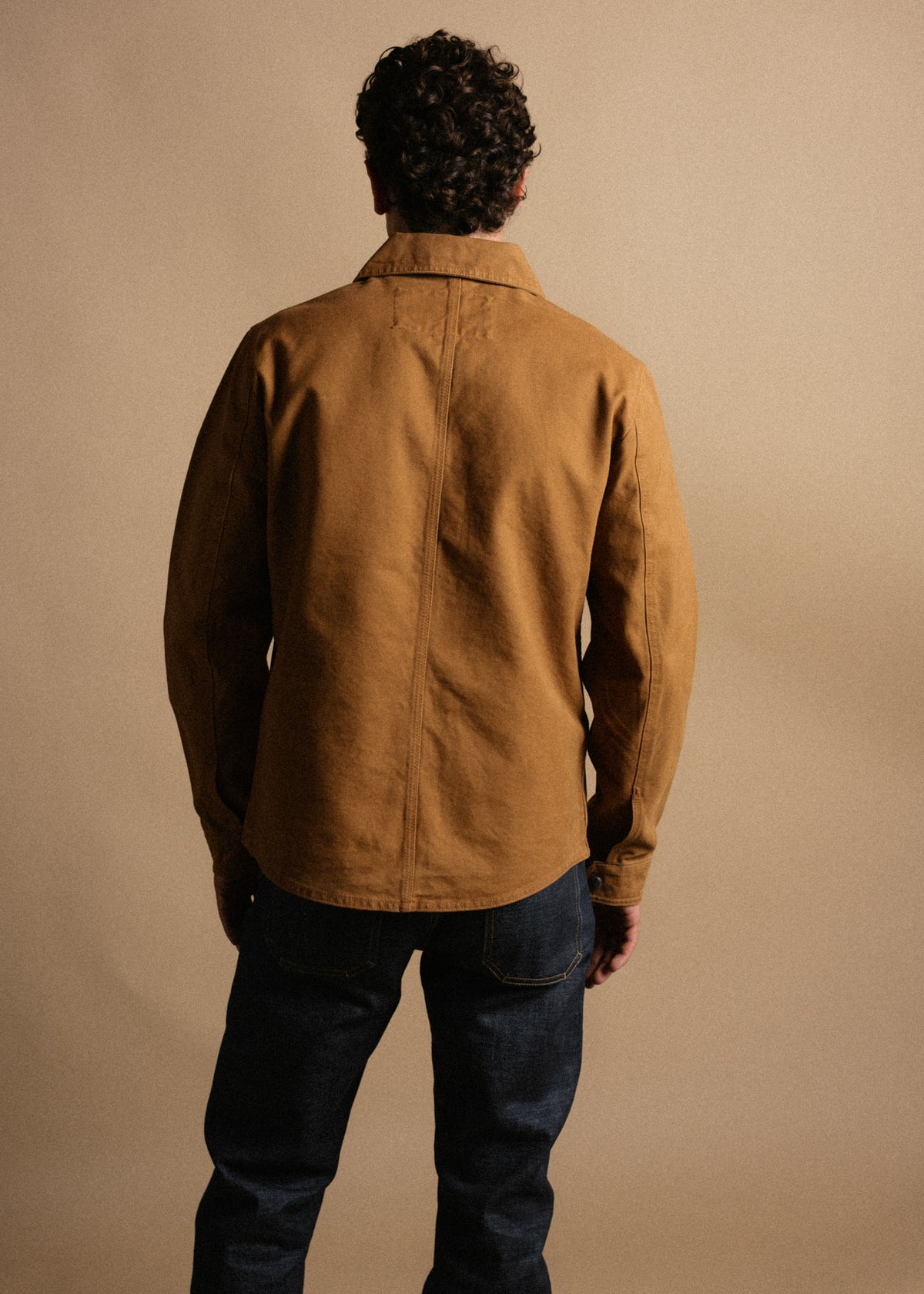Back view of a person wearing a brown jacket and dark jeans, standing against a beige background, highlighting casual style.