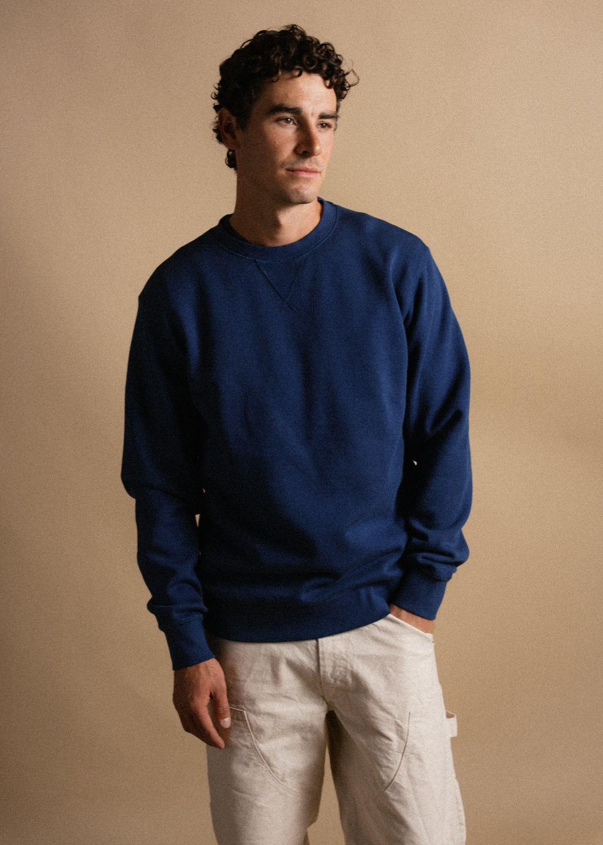 Man in a blue sweatshirt and beige pants stands against a neutral background, gazing thoughtfully to the side.
