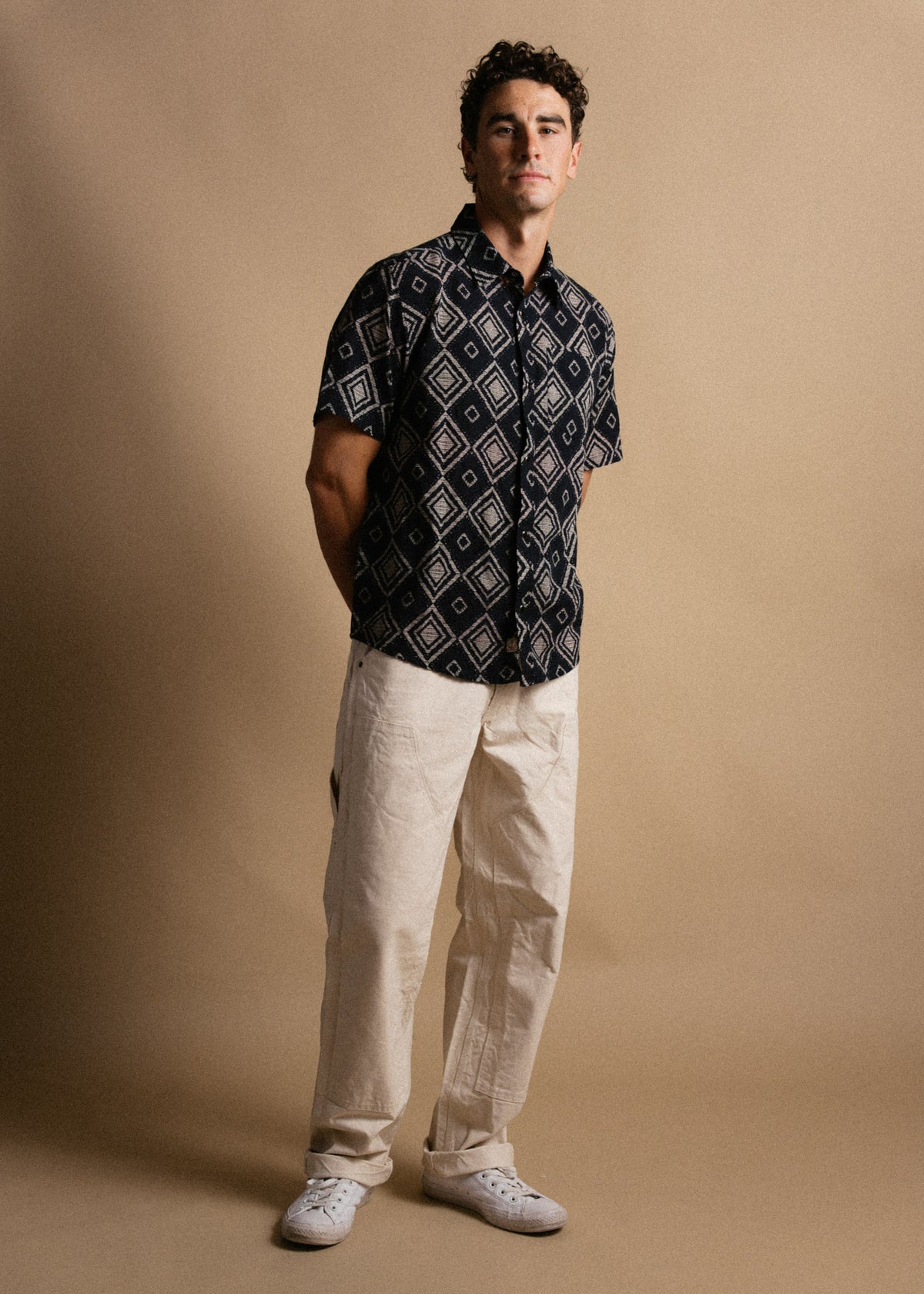 Man in a patterned shirt and beige pants stands against a plain background, hands behind back, exuding a casual, confident vibe.