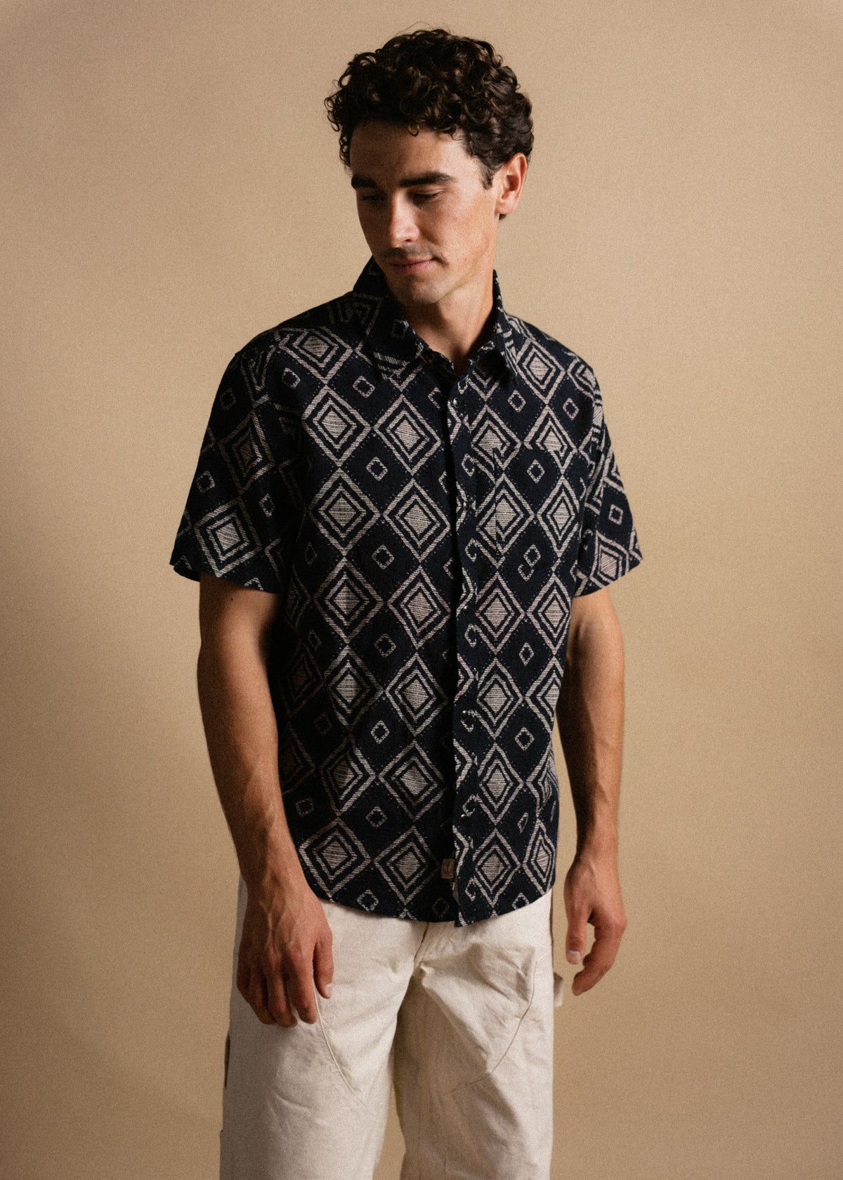 Man in a patterned shirt and white pants stands against a beige background, looking down with a thoughtful expression.
