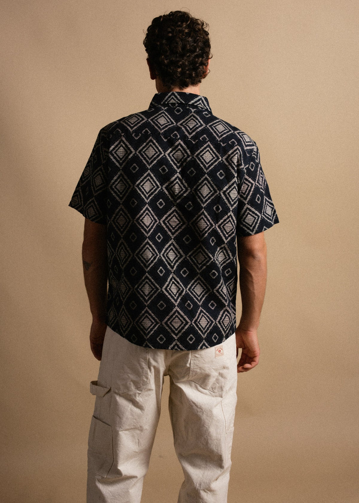 Man in a patterned navy shirt and white pants stands against a beige background, facing away, showcasing the clothing design.