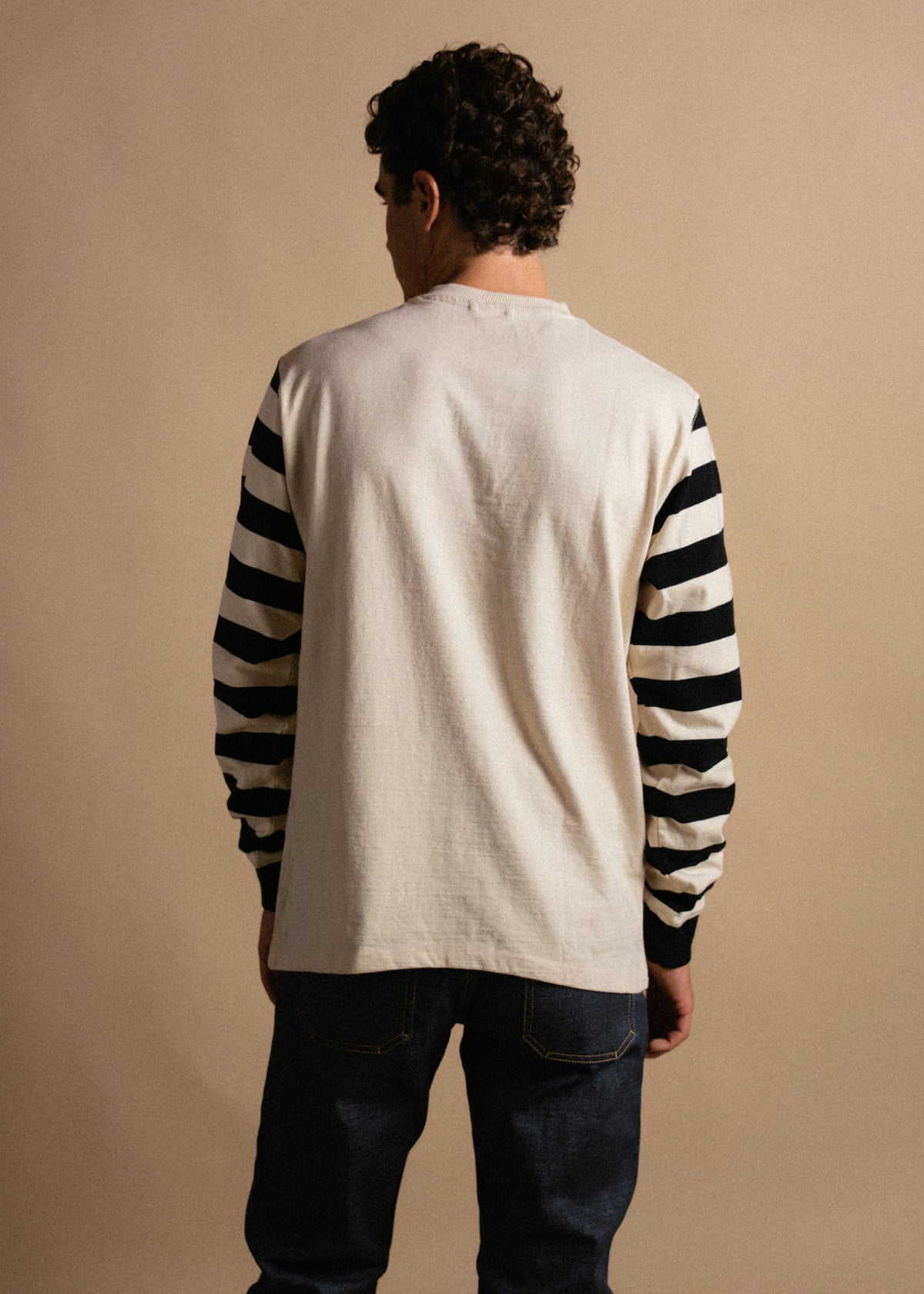 Man in a cream shirt with black-striped sleeves, facing away, stands against a beige background, wearing dark jeans.