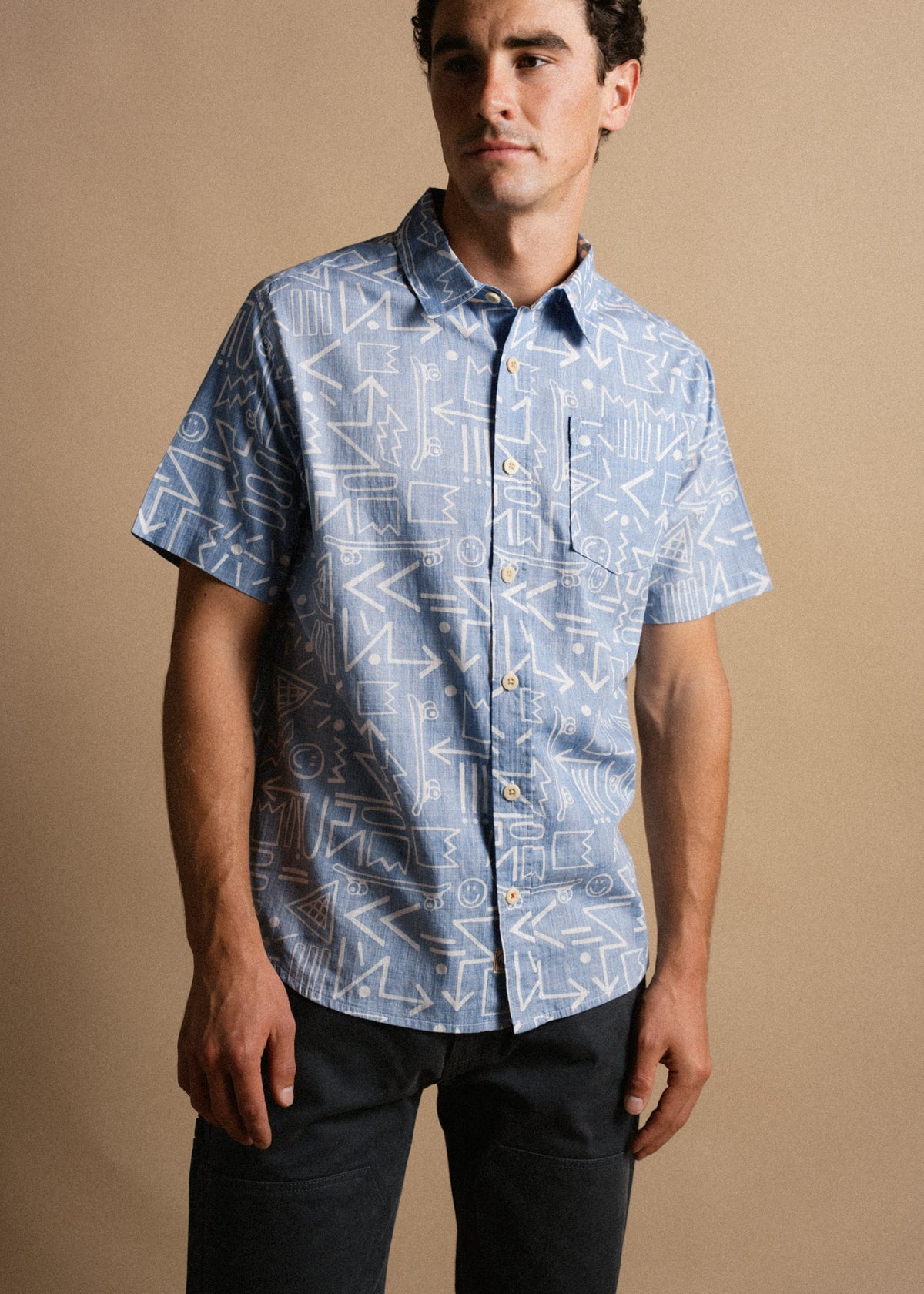 Man in a patterned blue shirt and dark pants stands against a beige background, looking to the side with a neutral expression.