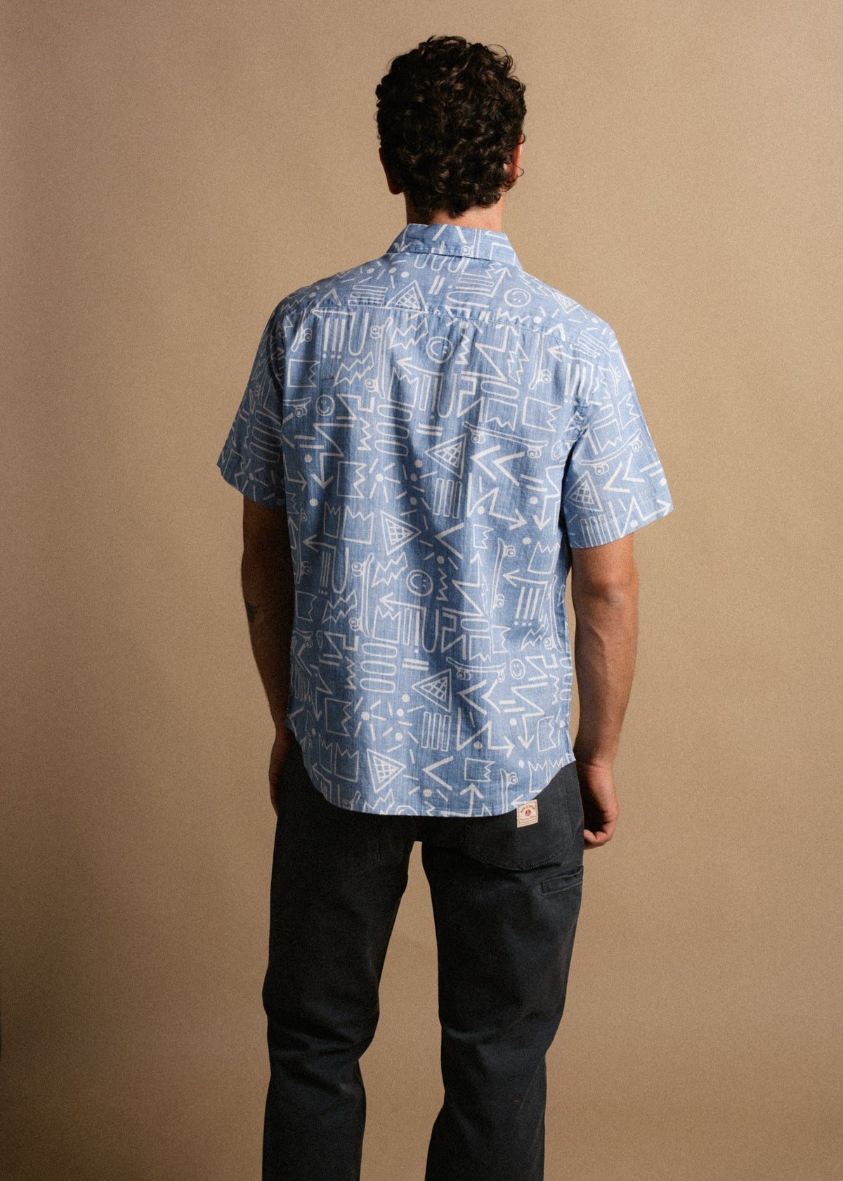 Man in blue patterned shirt and dark pants stands facing away against a beige background, showcasing the shirt's geometric design.
