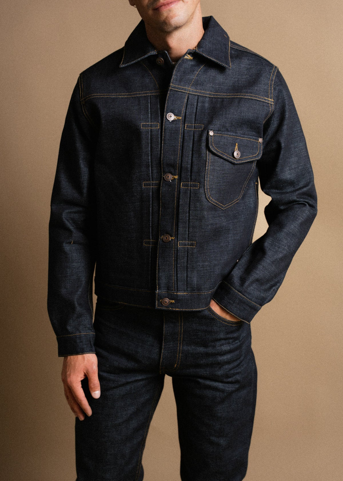 Man in dark denim jacket and jeans stands against a beige background, showcasing classic style and detailed stitching.