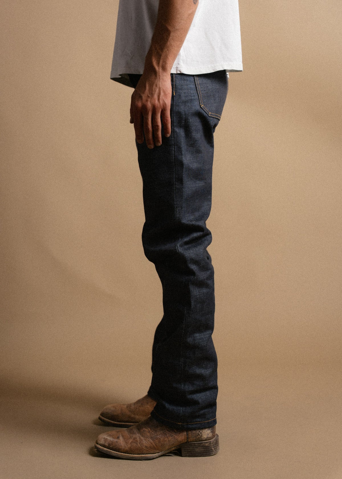 Man in a white shirt and dark jeans stands sideways, wearing worn brown boots, against a plain beige background.
