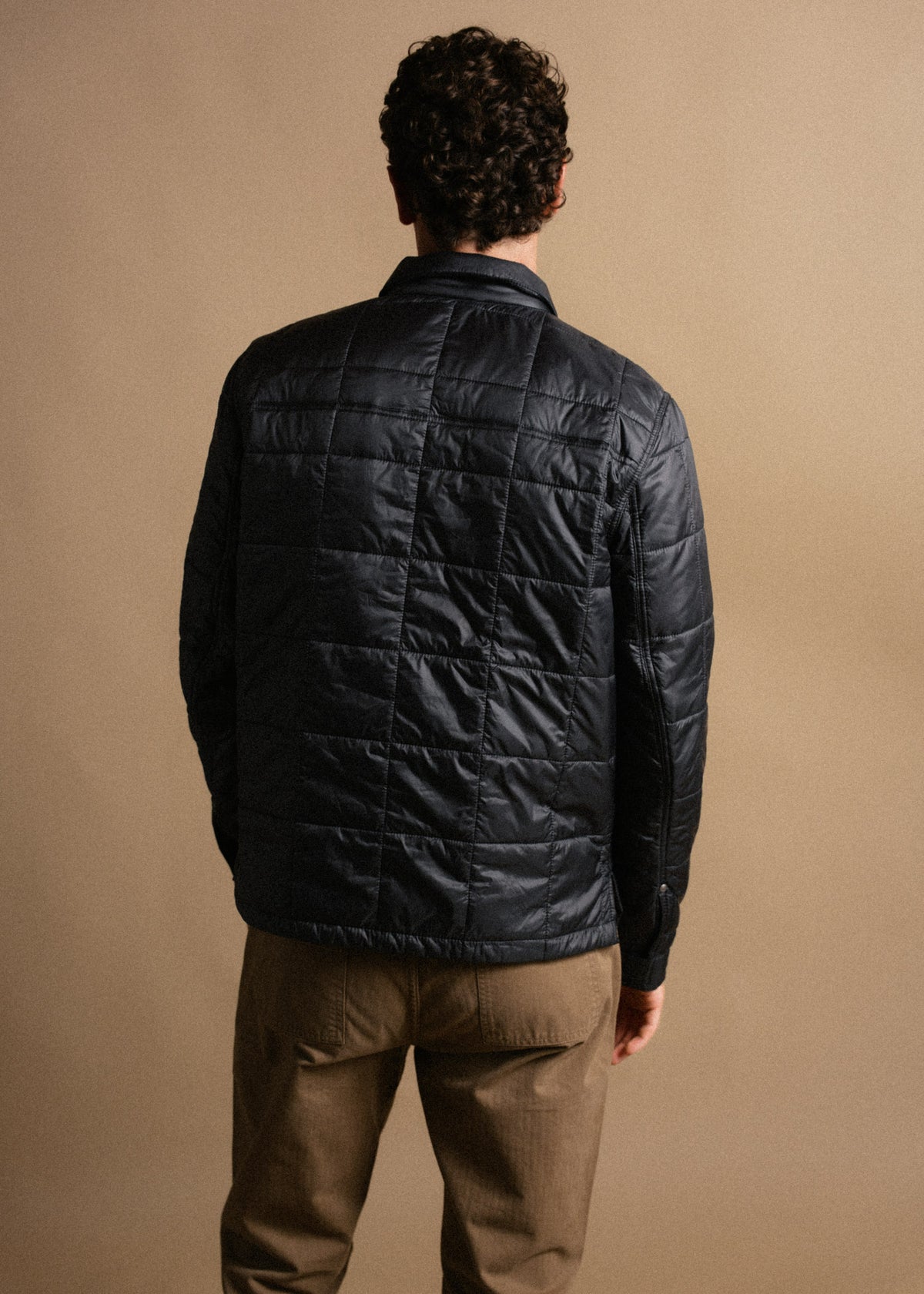 Man in a quilted navy jacket and brown pants stands with his back to the camera against a beige background, showcasing outerwear.