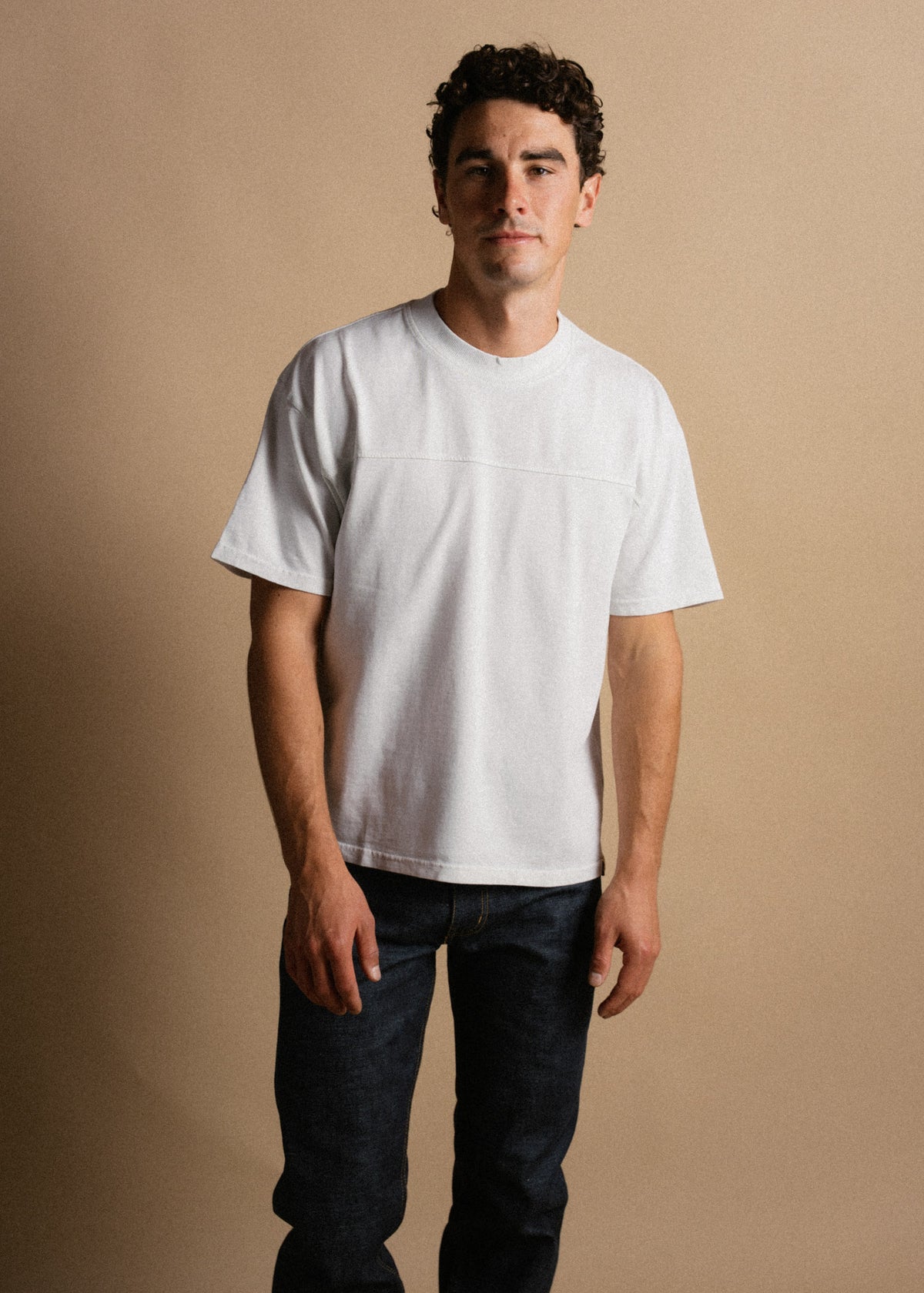Man in a white T-shirt and dark jeans stands against a beige background, looking confidently at the camera.