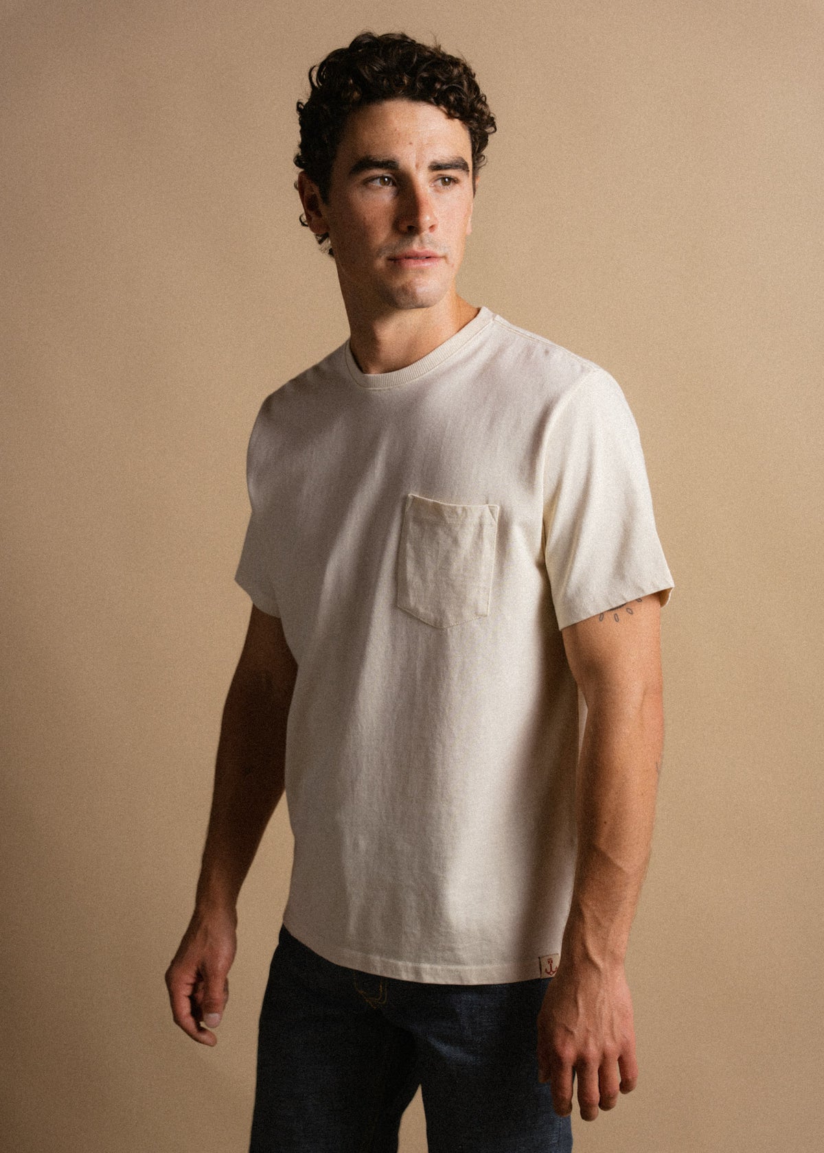Man in a white t-shirt with a pocket stands against a beige background, looking to the side with a thoughtful expression.