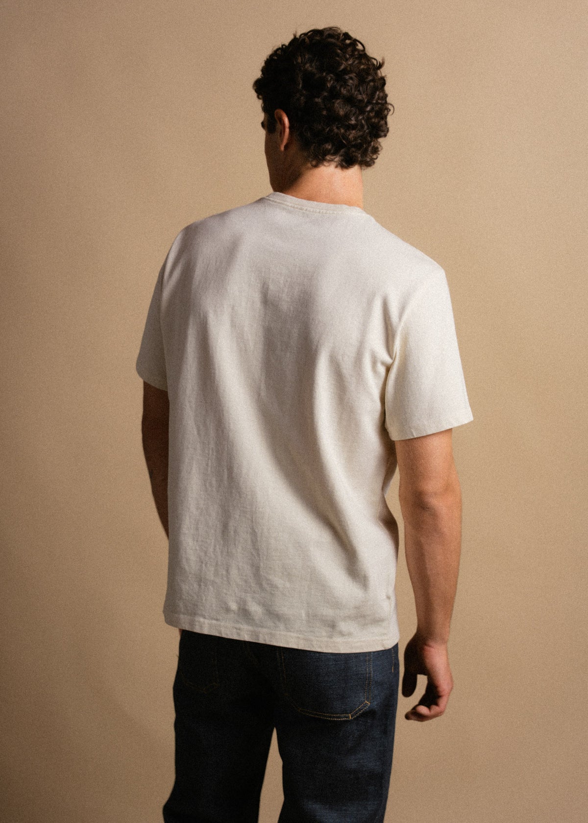 Man in a white t-shirt and jeans stands with his back to the camera against a beige background, showcasing casual style.