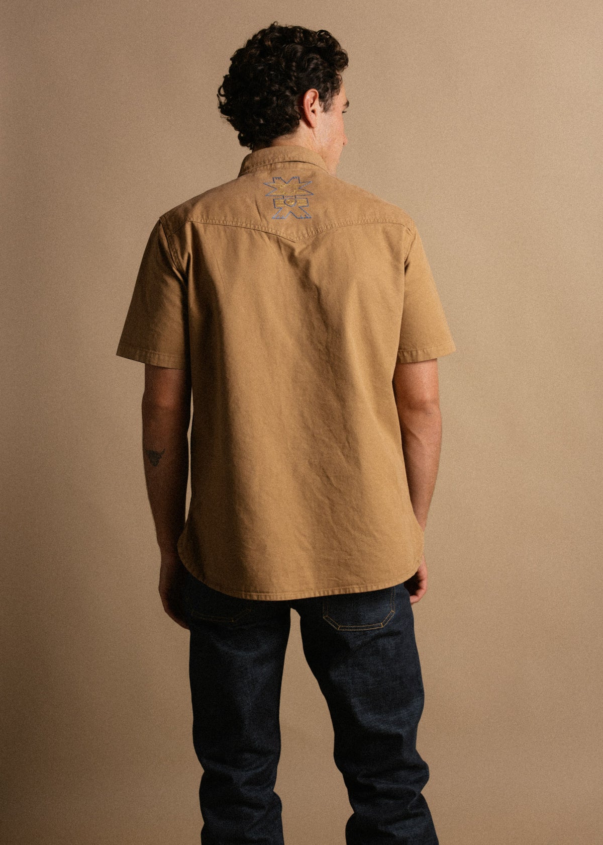 Man in a tan shirt with embroidered logo on back, facing away. Dark jeans, neutral background. Casual fashion style.