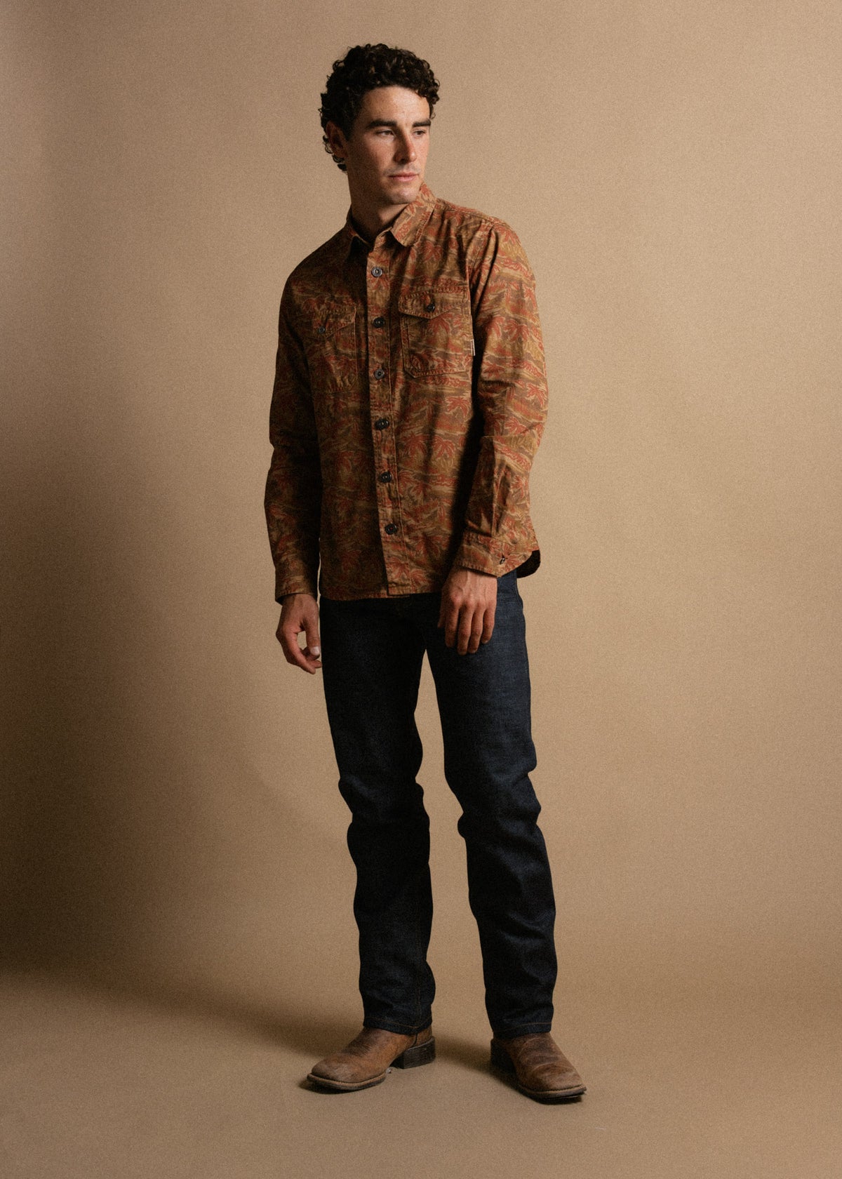 Man in a patterned brown shirt and jeans stands against a beige background, looking to the side with a thoughtful expression.