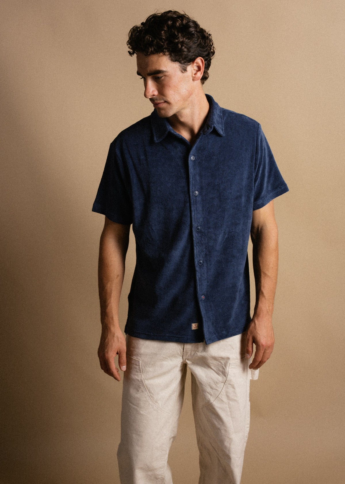 Man in a navy blue button-up shirt and beige pants stands against a beige background, looking down to his left.