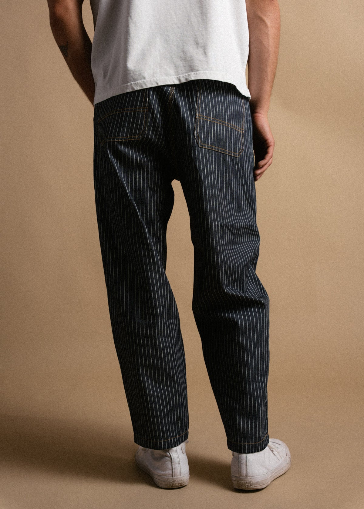 Person in white shirt and pinstriped pants stands against a beige background, showcasing casual style with white sneakers.
