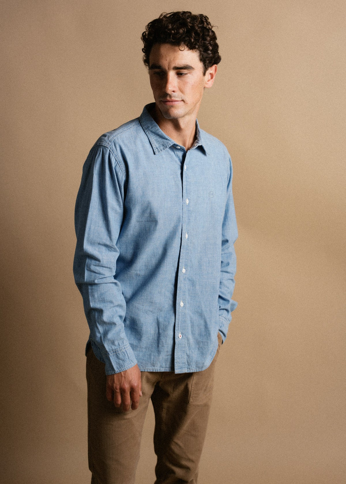 A man in a blue shirt and brown pants stands against a beige background, looking down thoughtfully with a neutral expression.