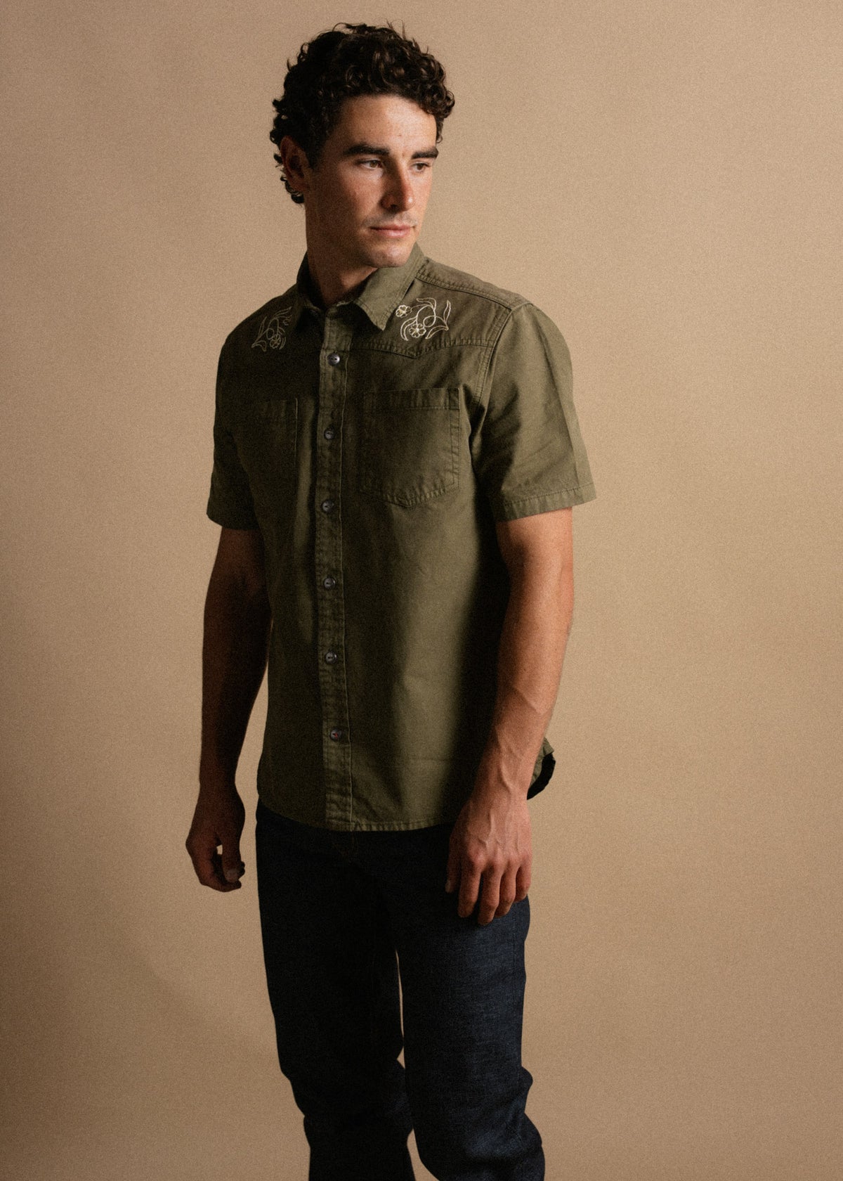 Man in olive shirt with embroidered details stands against a beige background, looking to the side. Casual and stylish attire.