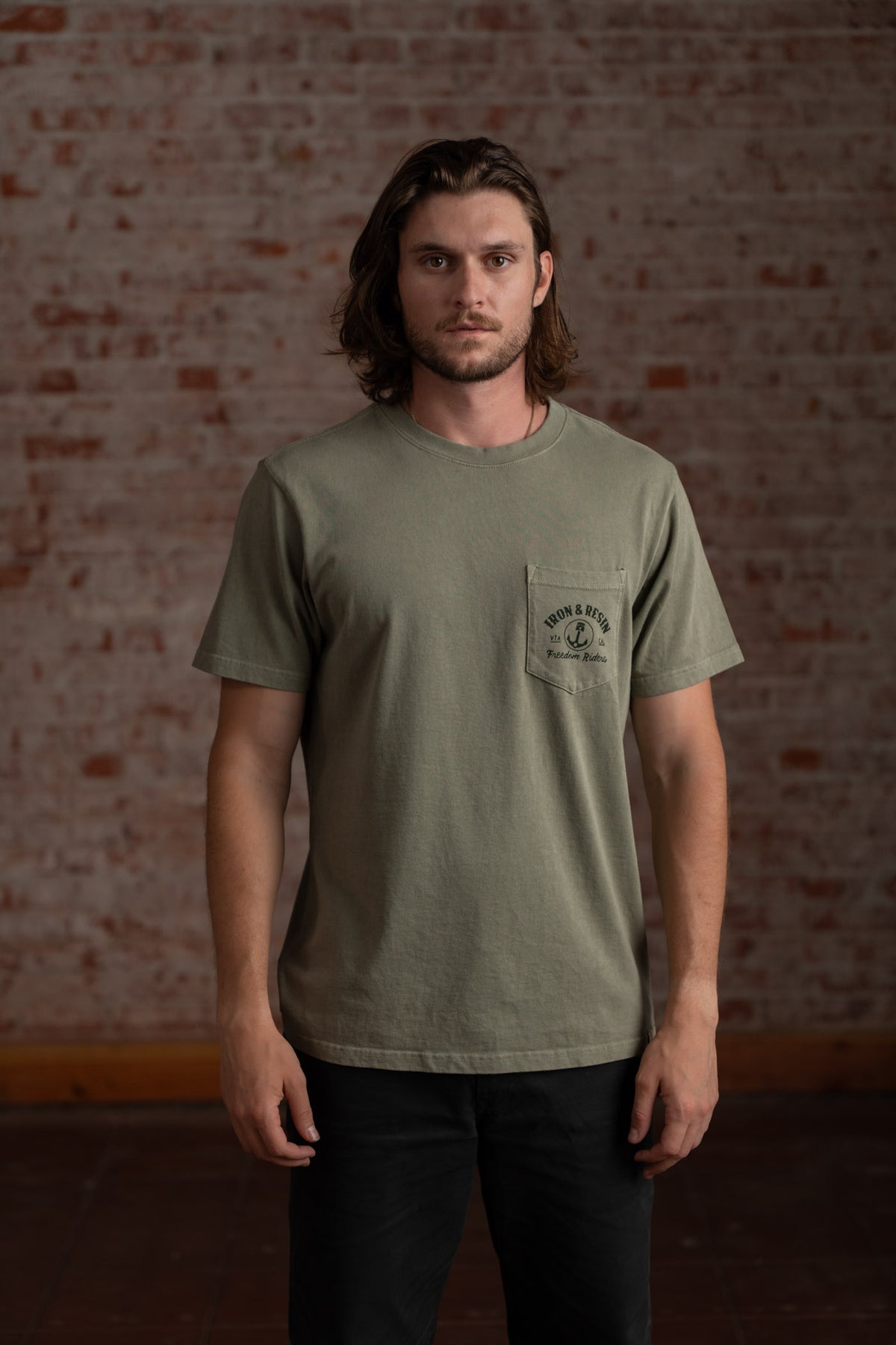 Man in green t-shirt with logo stands against a textured brick wall, looking forward. Casual style, relaxed pose, indoor setting.
