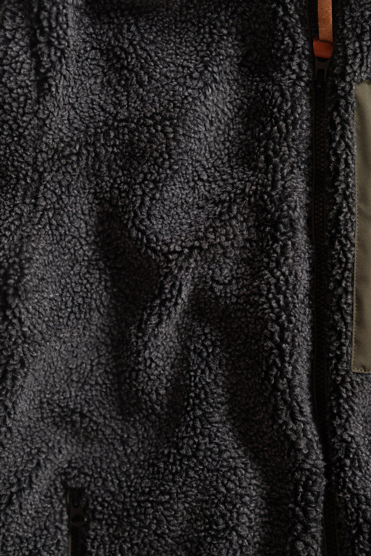 Close-up of a dark gray fleece jacket texture with visible zipper and brown leather pull tab, highlighting its cozy material.