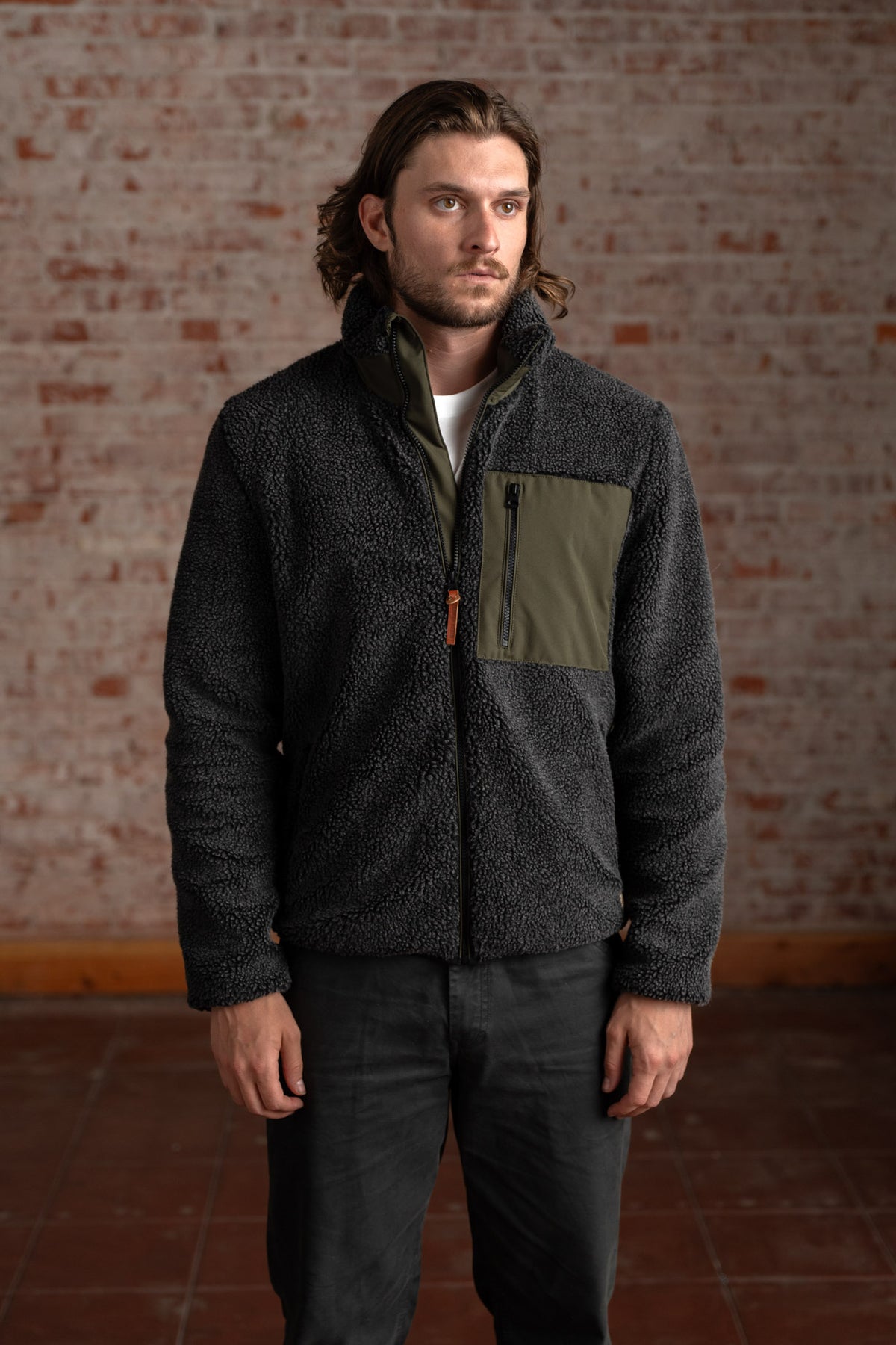 Man in a cozy black fleece jacket with green pocket stands against a brick wall, looking to the side, in a casual setting.