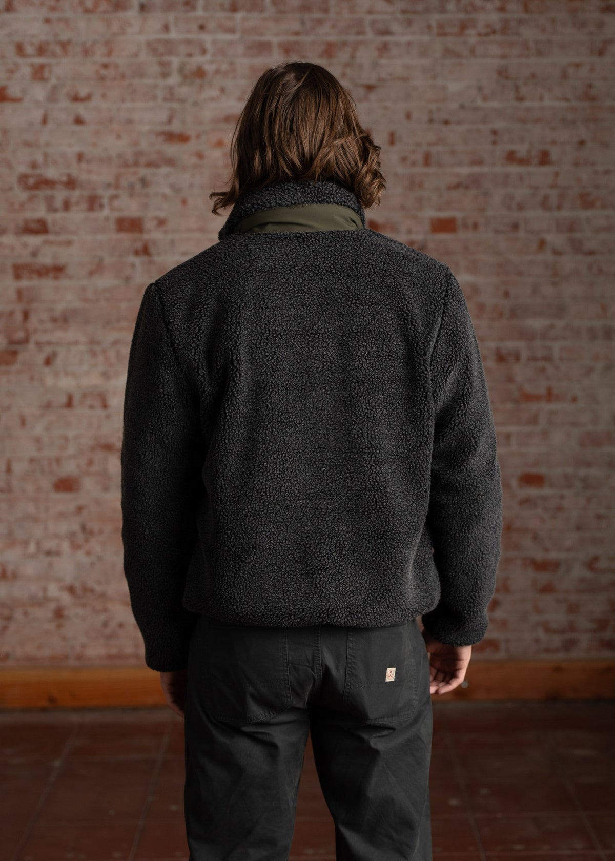 Man in a black fleece jacket and dark pants stands facing a brick wall, highlighting the textured fabric and casual style.