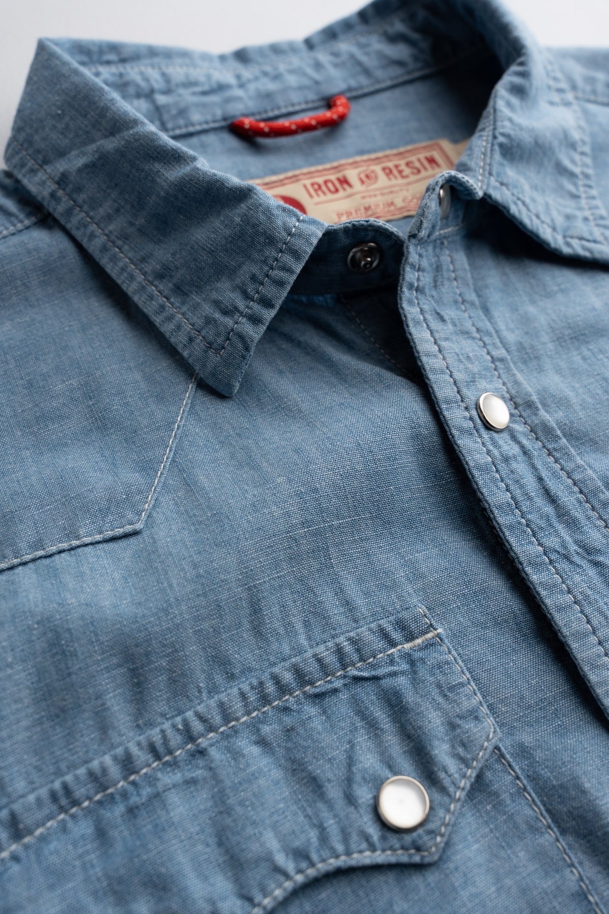Close up of woven neck label and interior locker loop on chambray shirt, highlighting pearl snaps and heritage workwear details