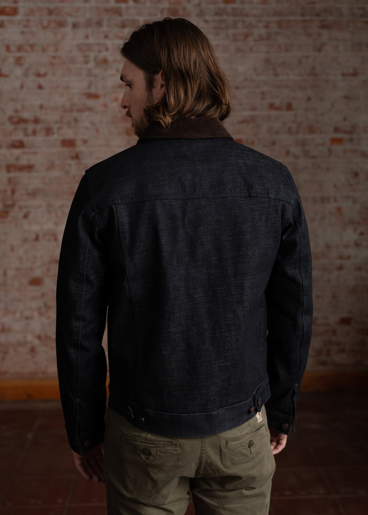 Man with long hair in a dark denim jacket stands against a textured brick wall, facing away, in a casual indoor setting.