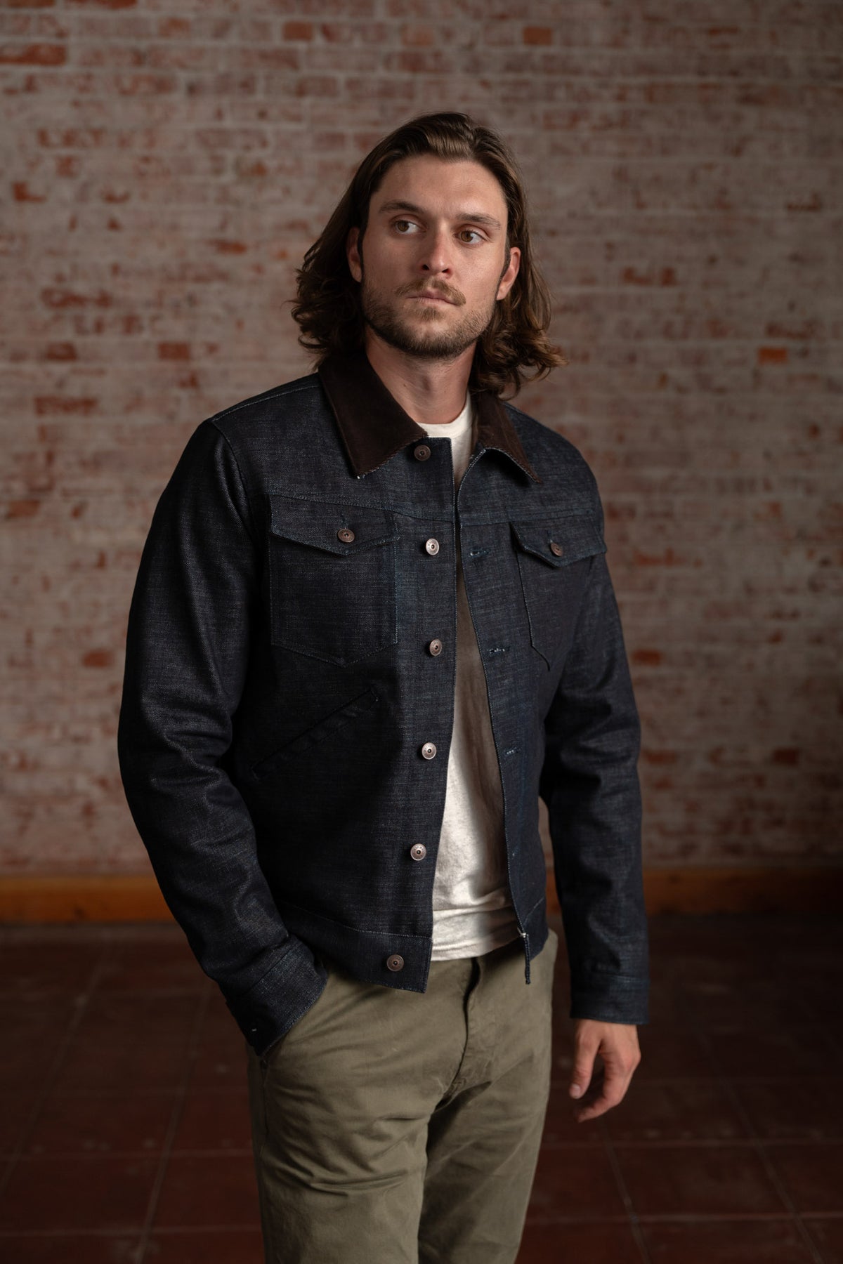 Man in a denim jacket with a brown collar stands against a brick wall, looking to the side, wearing khaki pants and a white shirt.