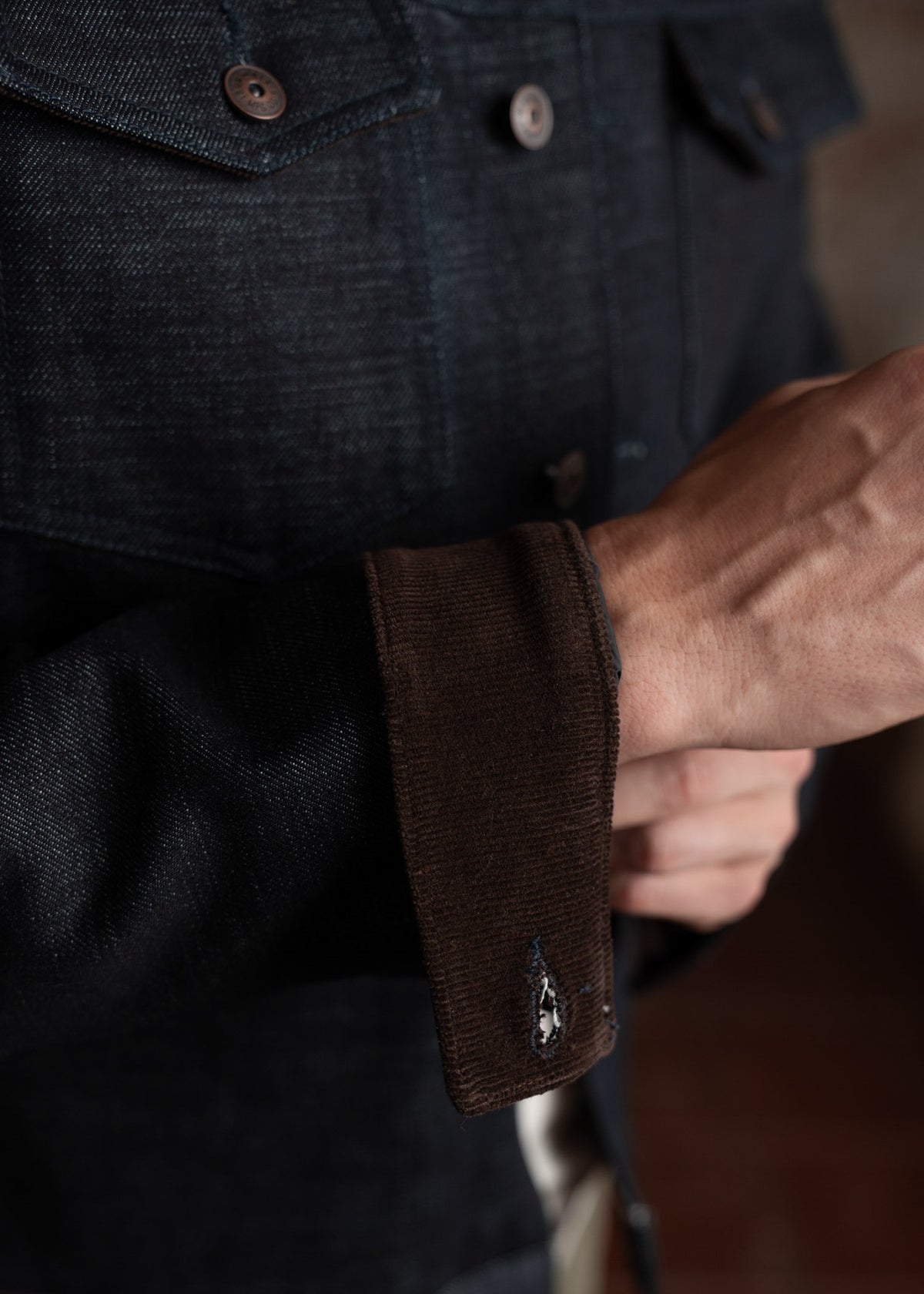 Close-up of a person adjusting a dark denim jacket sleeve, highlighting the button details and texture in a casual setting.