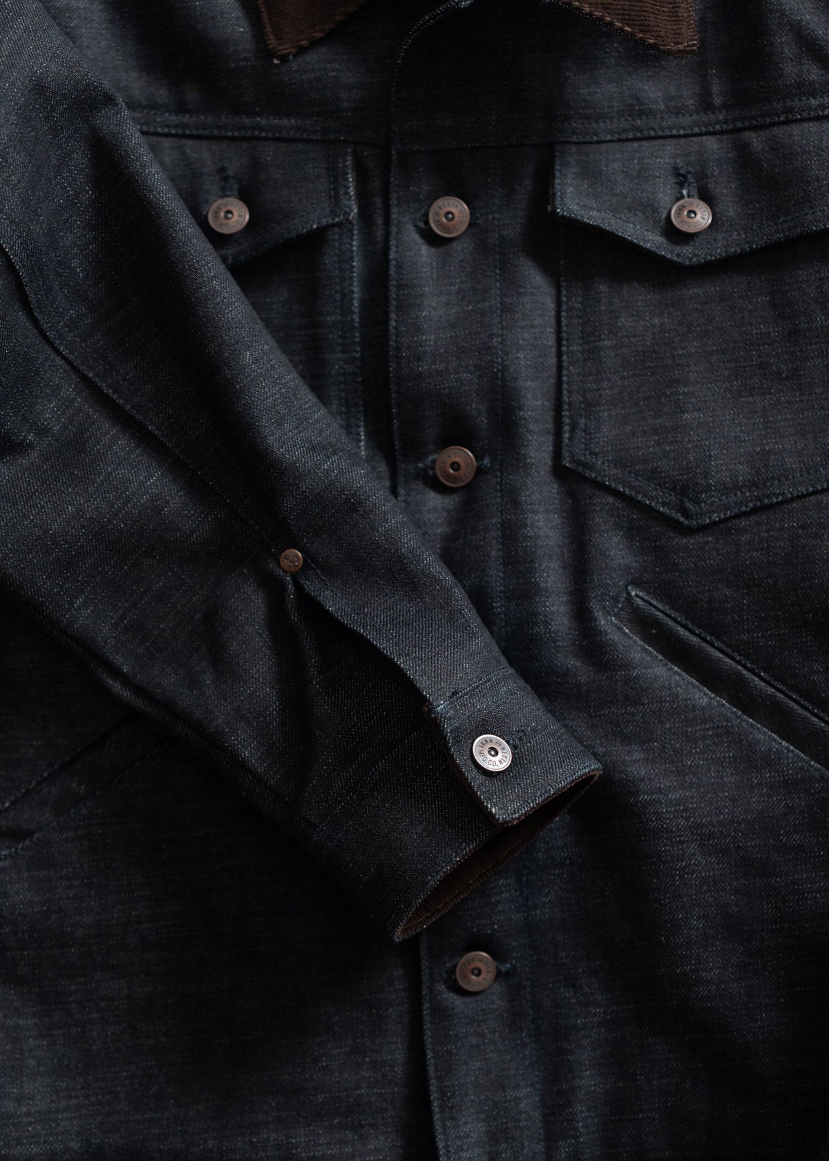 Close-up of a dark denim jacket with buttoned pockets and sleeves, highlighting texture and stitching details.