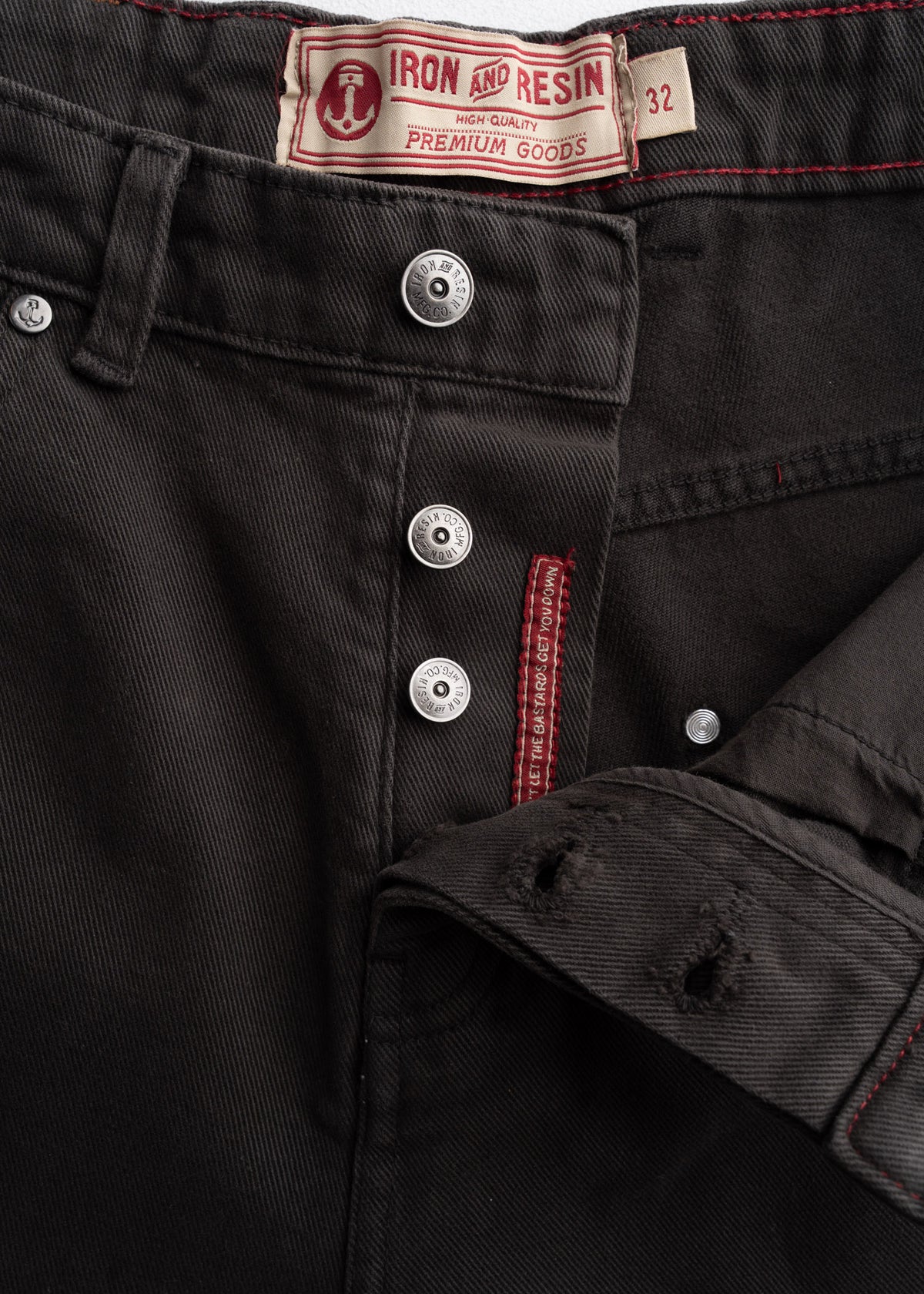 Close-up of Iron and Resin black jeans with red stitching, button fly, and label showing size 32, emphasizing premium quality.