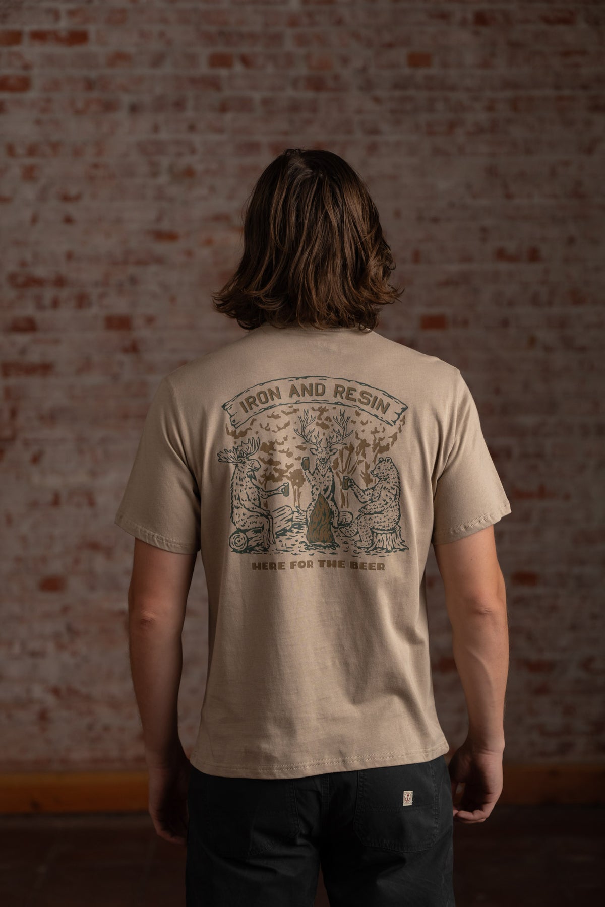 Man in beige "Iron and Resin" T-shirt with forest scene and "Here for the Beer" text, standing against a brick wall.