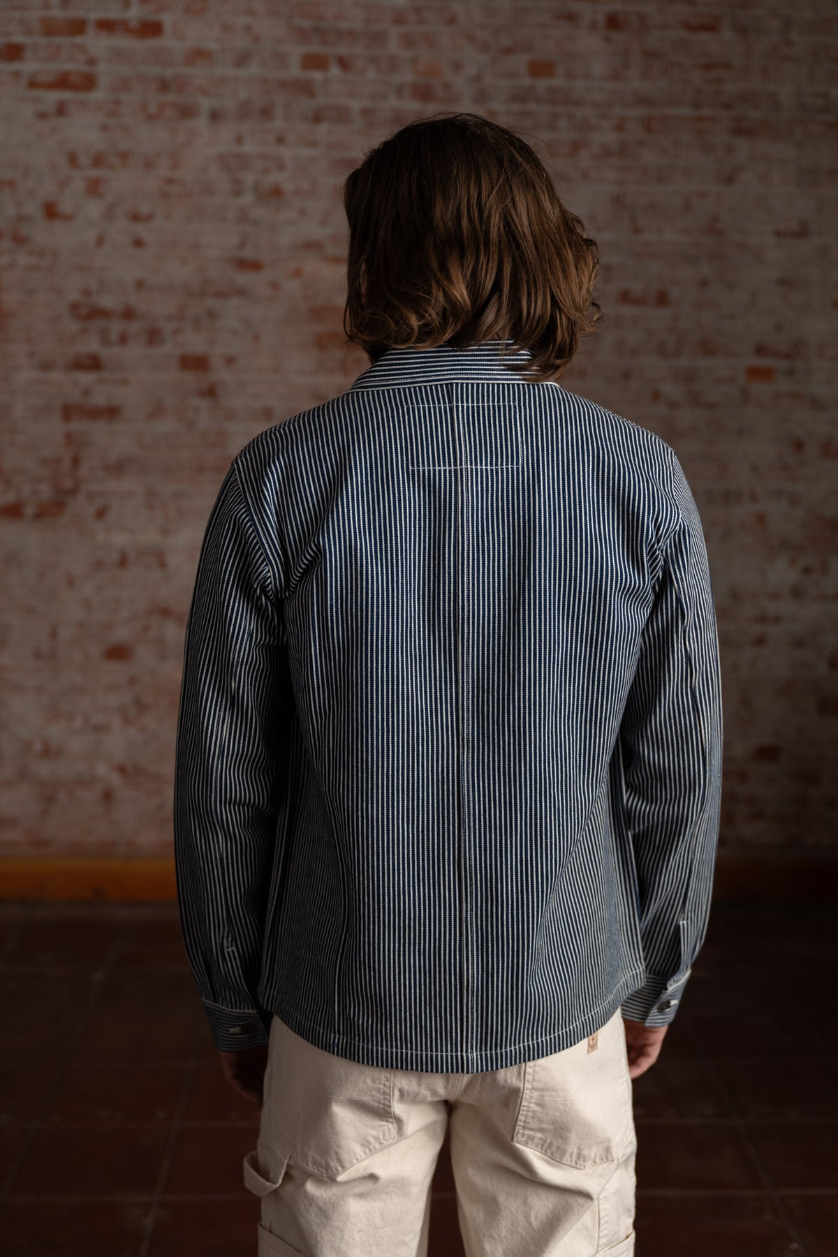 Person with shoulder-length hair in a striped shirt and beige pants stands against a brick wall, facing away.