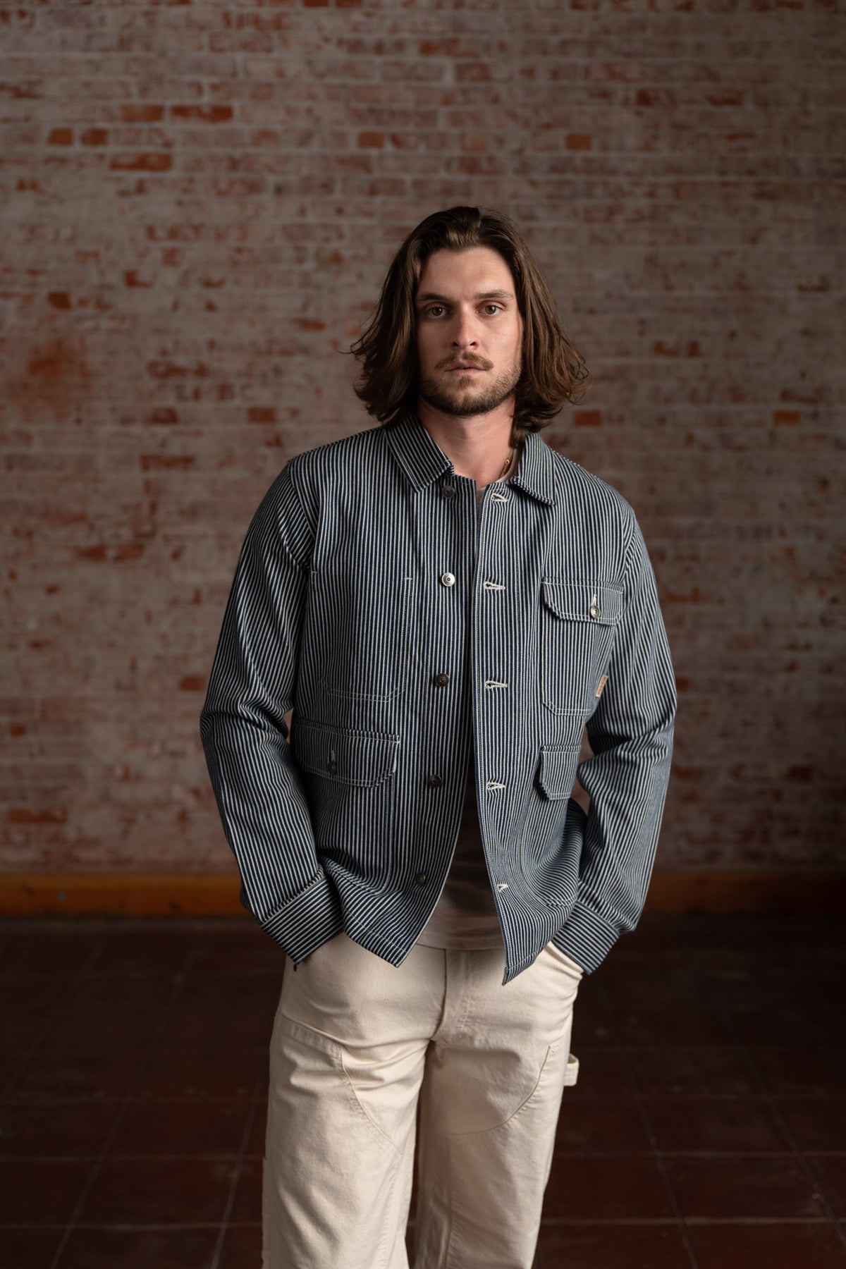A man with long hair wears a striped jacket and beige pants, standing against a textured brick wall, hands in pockets.